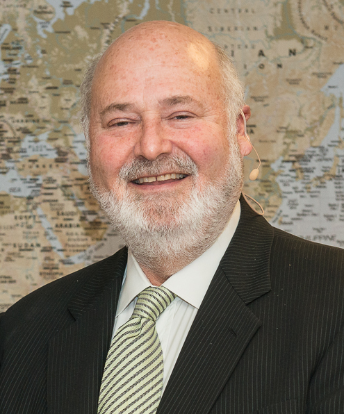 Smiling bald man with white beard in dark suit and green striped tie at a public event.