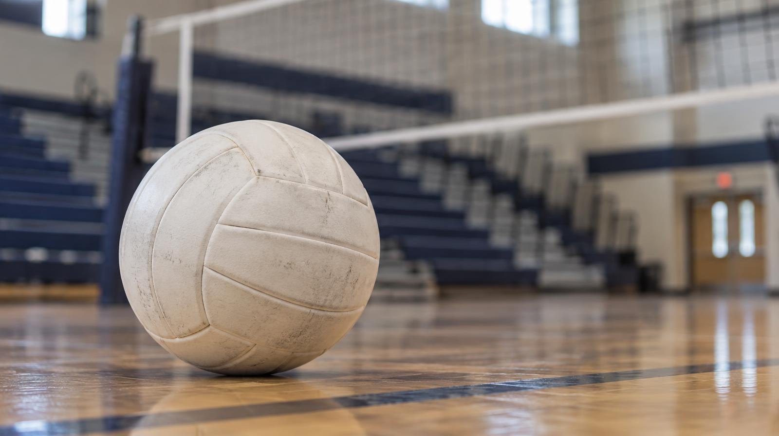 A white volleyball resting on a hardwood gymnasium floor near the net.