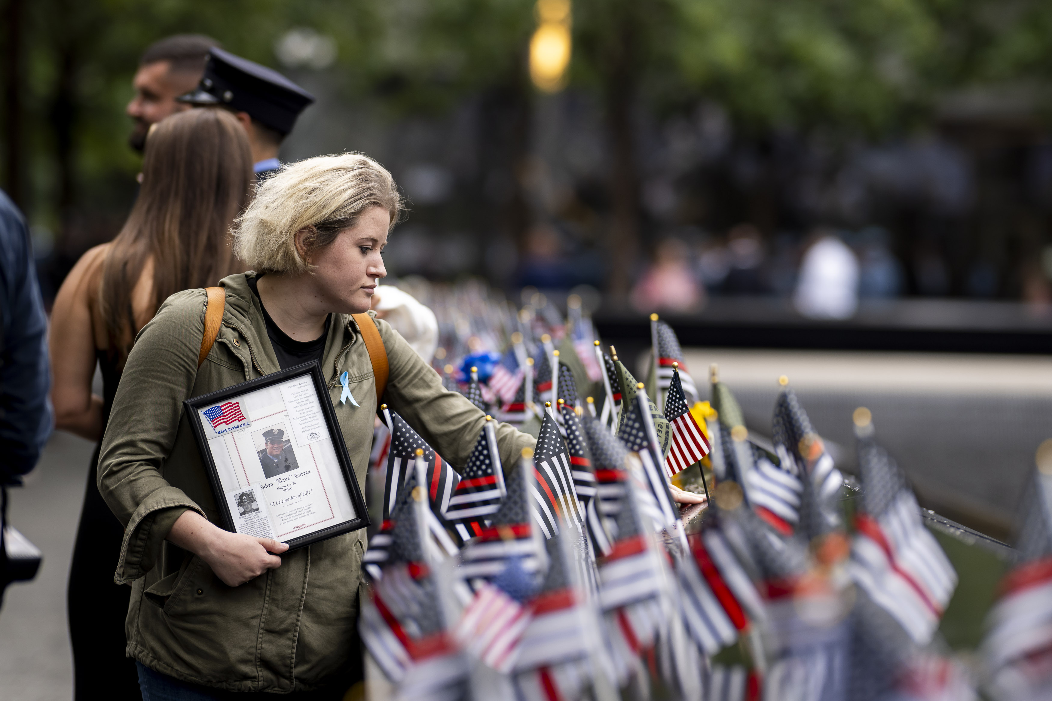 Woman holding a framed photo among rows of small flags at a memorial reflecting pool.