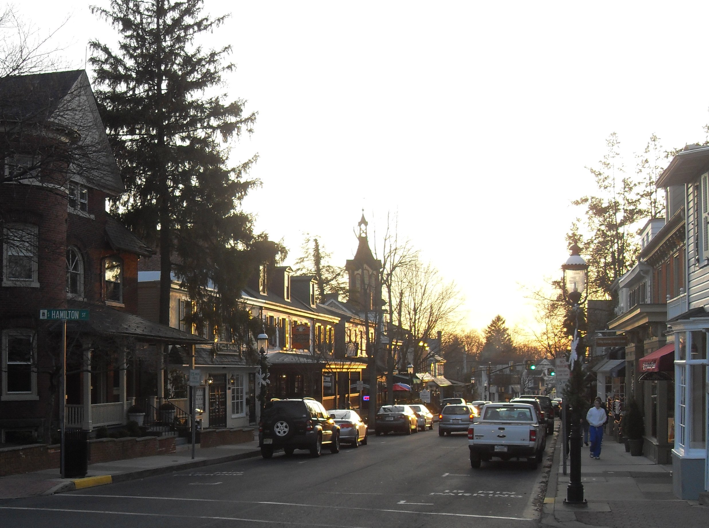 Brick commercial buildings line a small-town main street at dusk with a clock tower in the background.