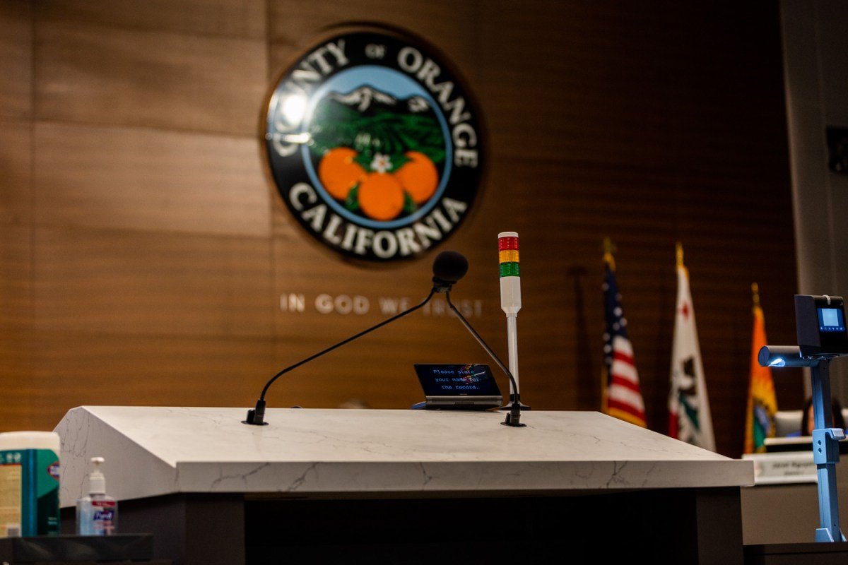 An empty podium with a microphone and speaker timer in front of the County of Orange seal and flags.
