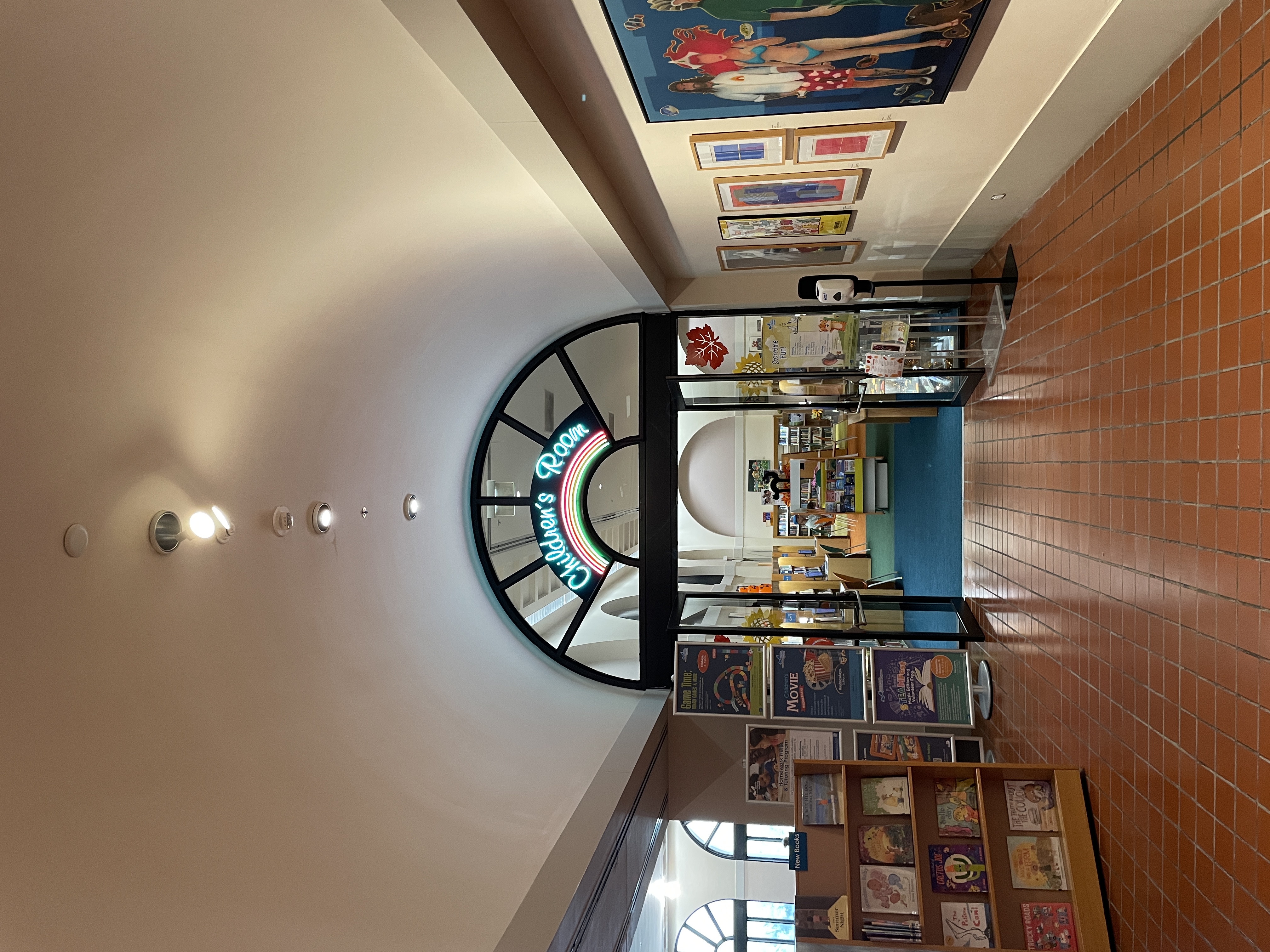 Arched library corridor leading to a children's room entrance with a neon rainbow sign.