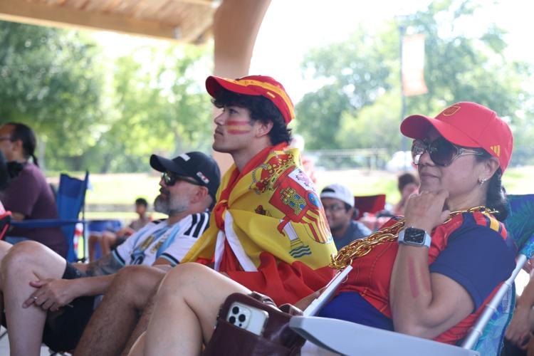 Two fans in red Spain gear, one draped in a Spanish flag, watch an outdoor event.