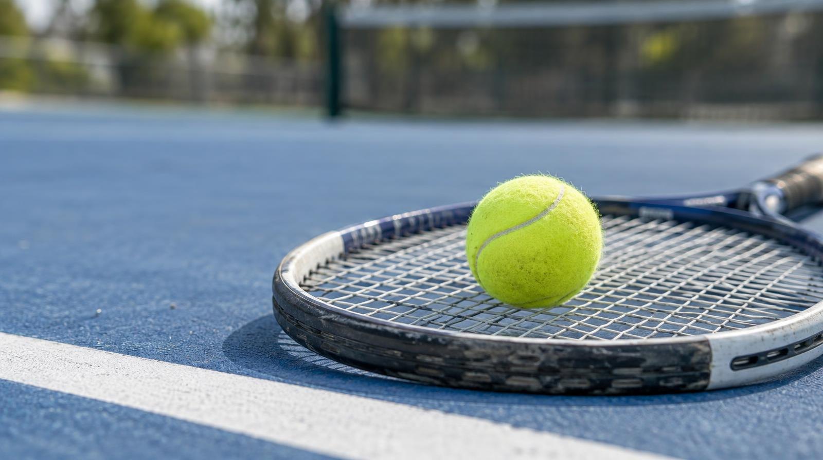Yellow tennis ball resting on the strings of a racket on a blue hard court.