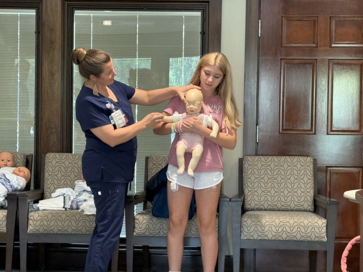 Instructor in blue scrubs helps a teenage girl hold a baby mannequin indoors.