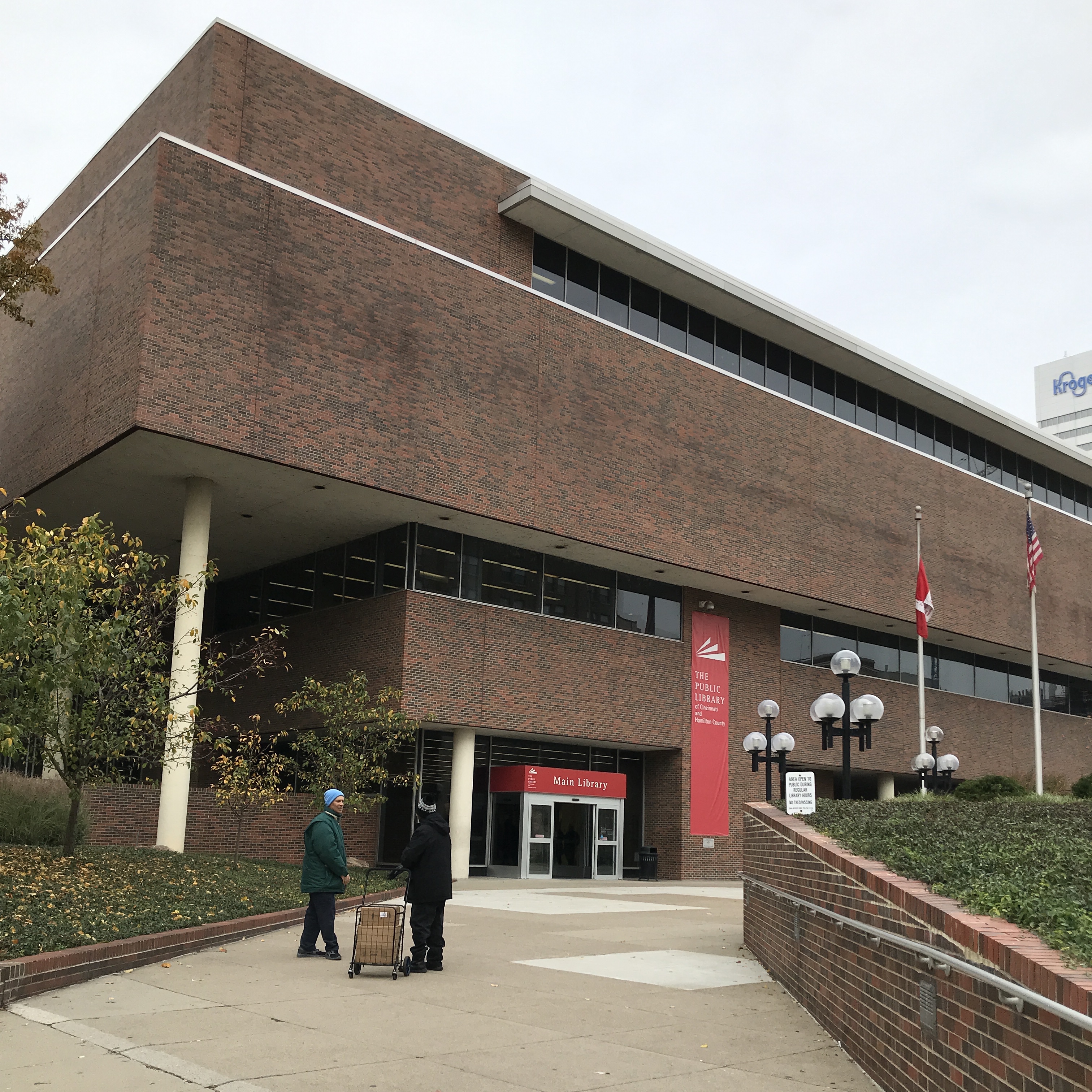 Exterior of a multi-story brick library building with flagpoles and a red banner sign.