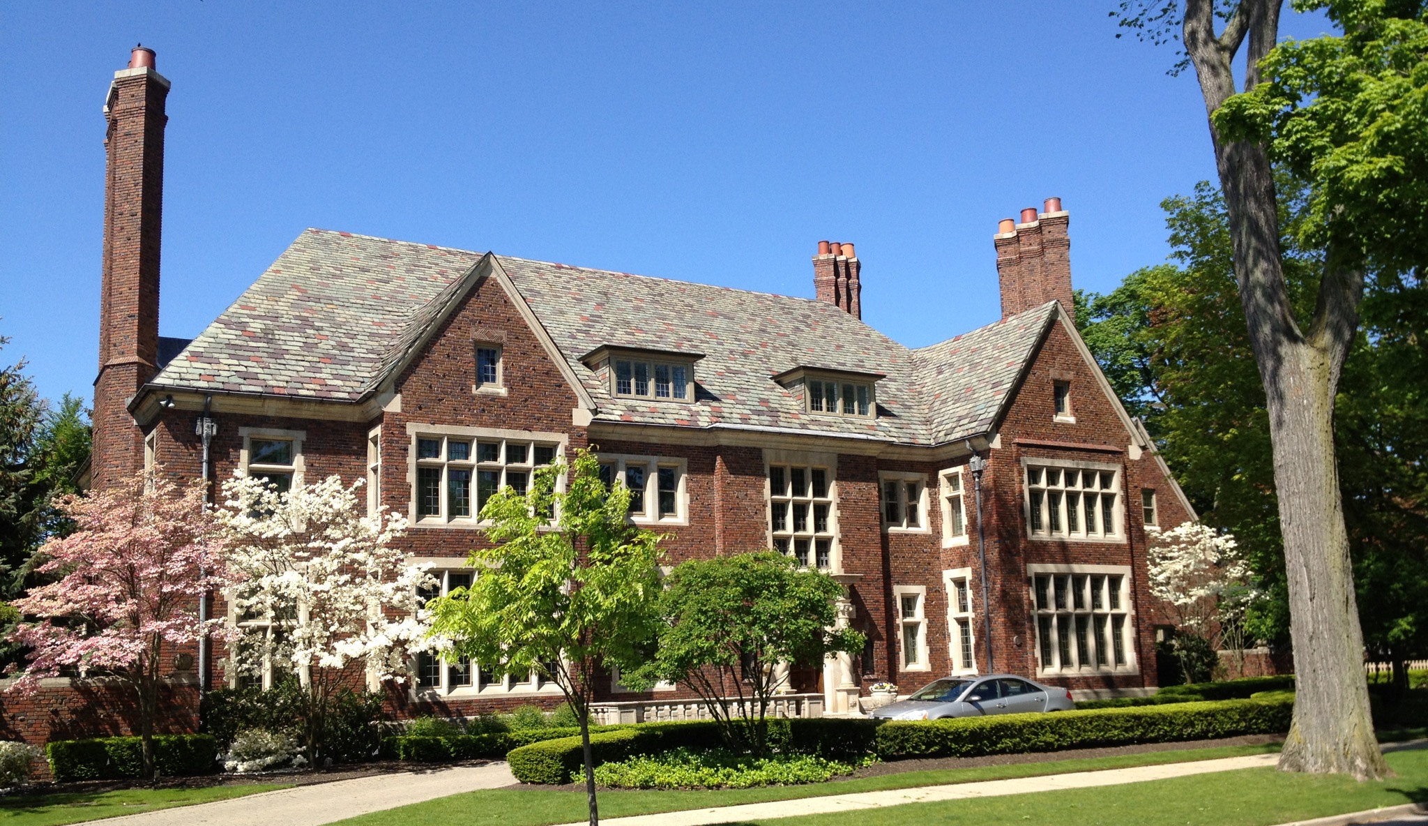 Large Tudor-style brick mansion with slate roof, chimneys, and flowering trees on a sunny day.