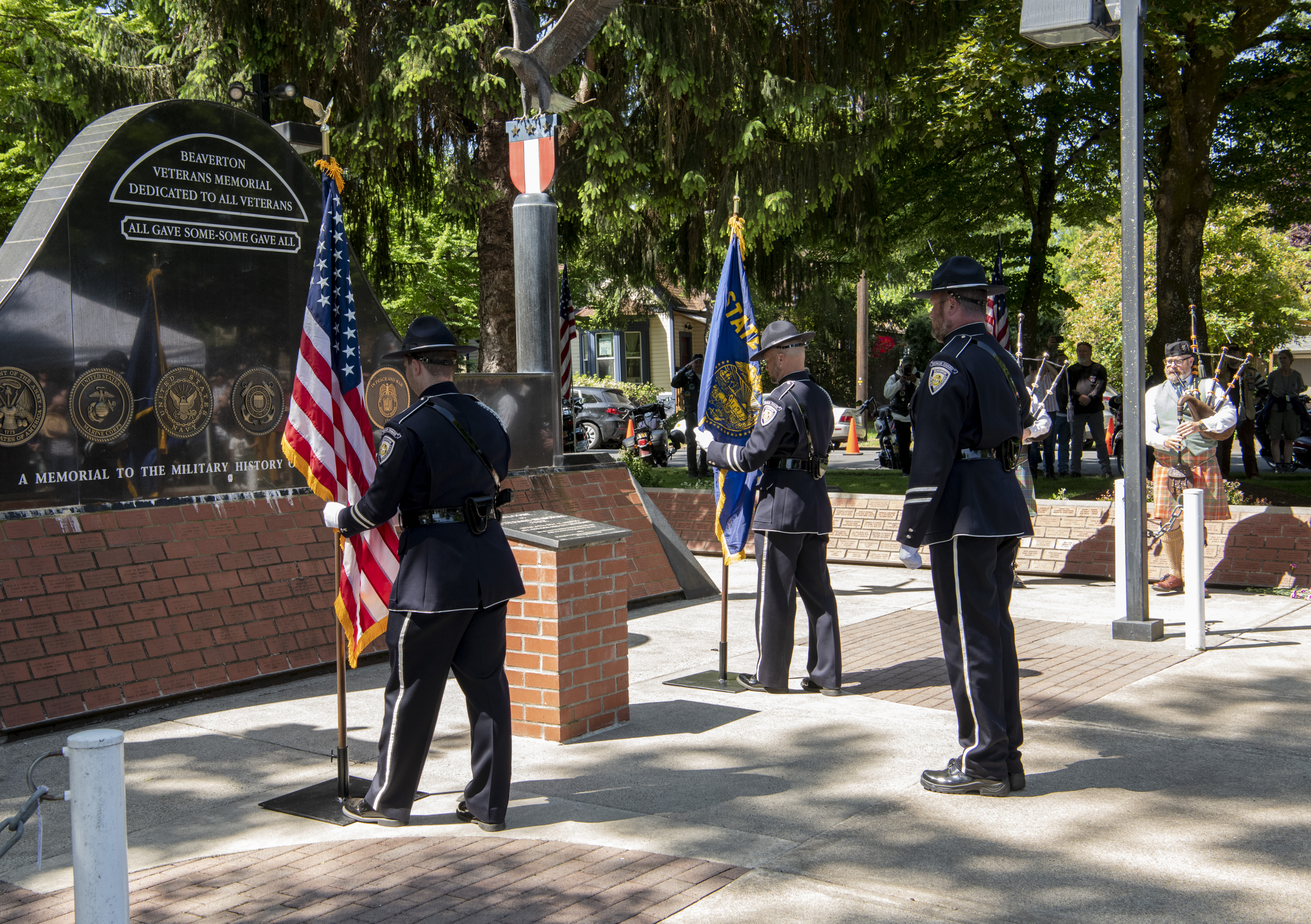 Three uniformed officers in a color guard holding flags at an outdoor veterans memorial.