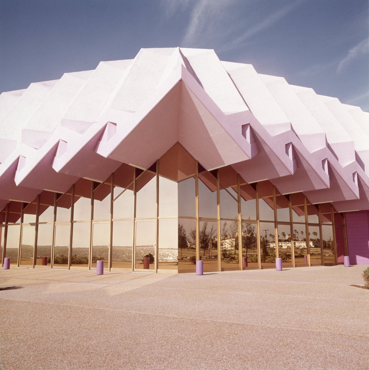 Pink folded-plate roof building with glass walls and purple bollards under blue sky.