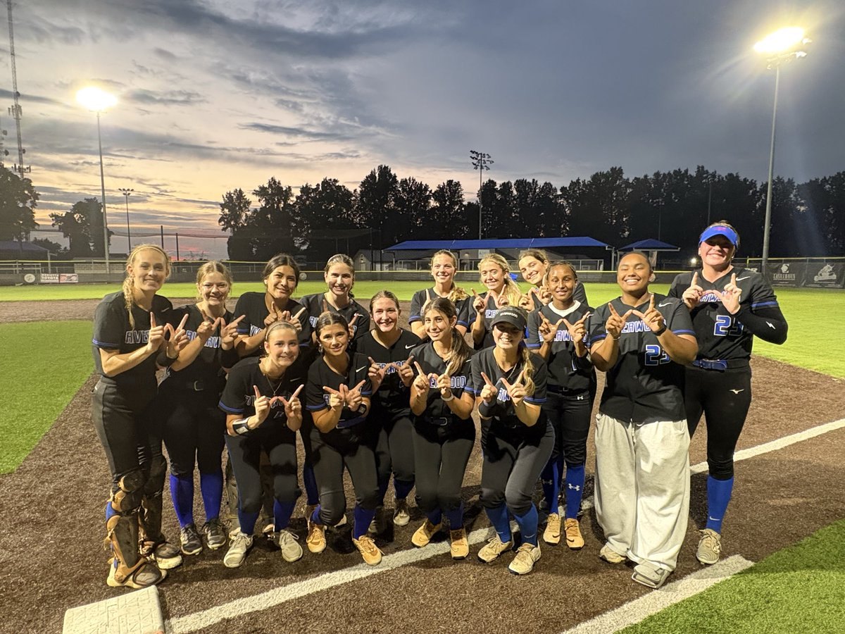 About 15 softball players in dark uniforms pose together on a field under stadium lights at dusk.