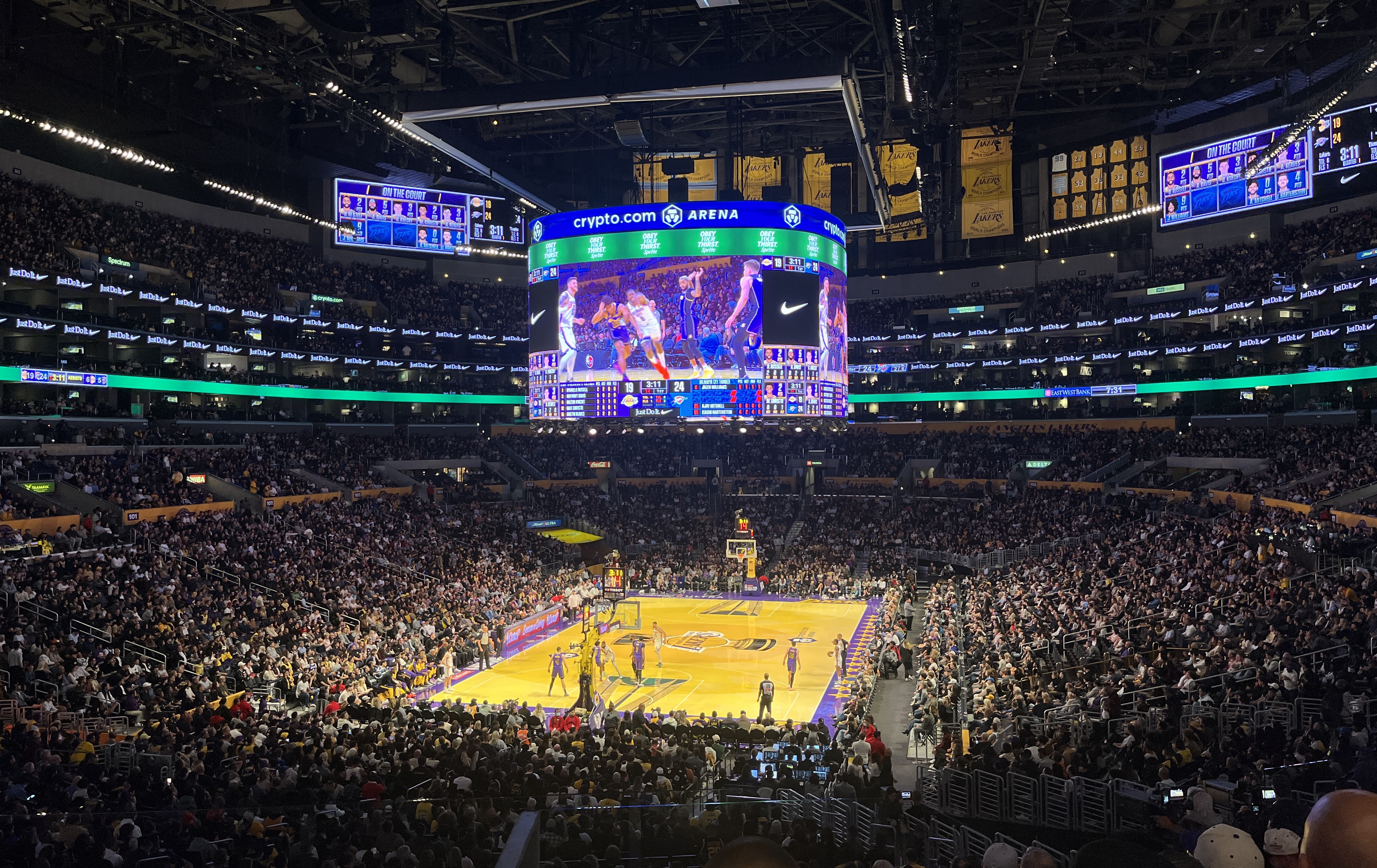 Packed NBA arena interior with purple and gold Lakers court and center-hung scoreboard.
