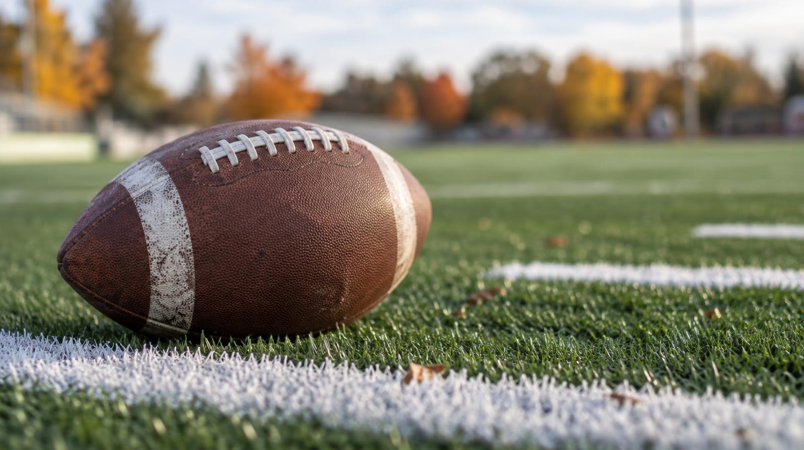 An American football resting on green artificial turf beside white yard-line markings.