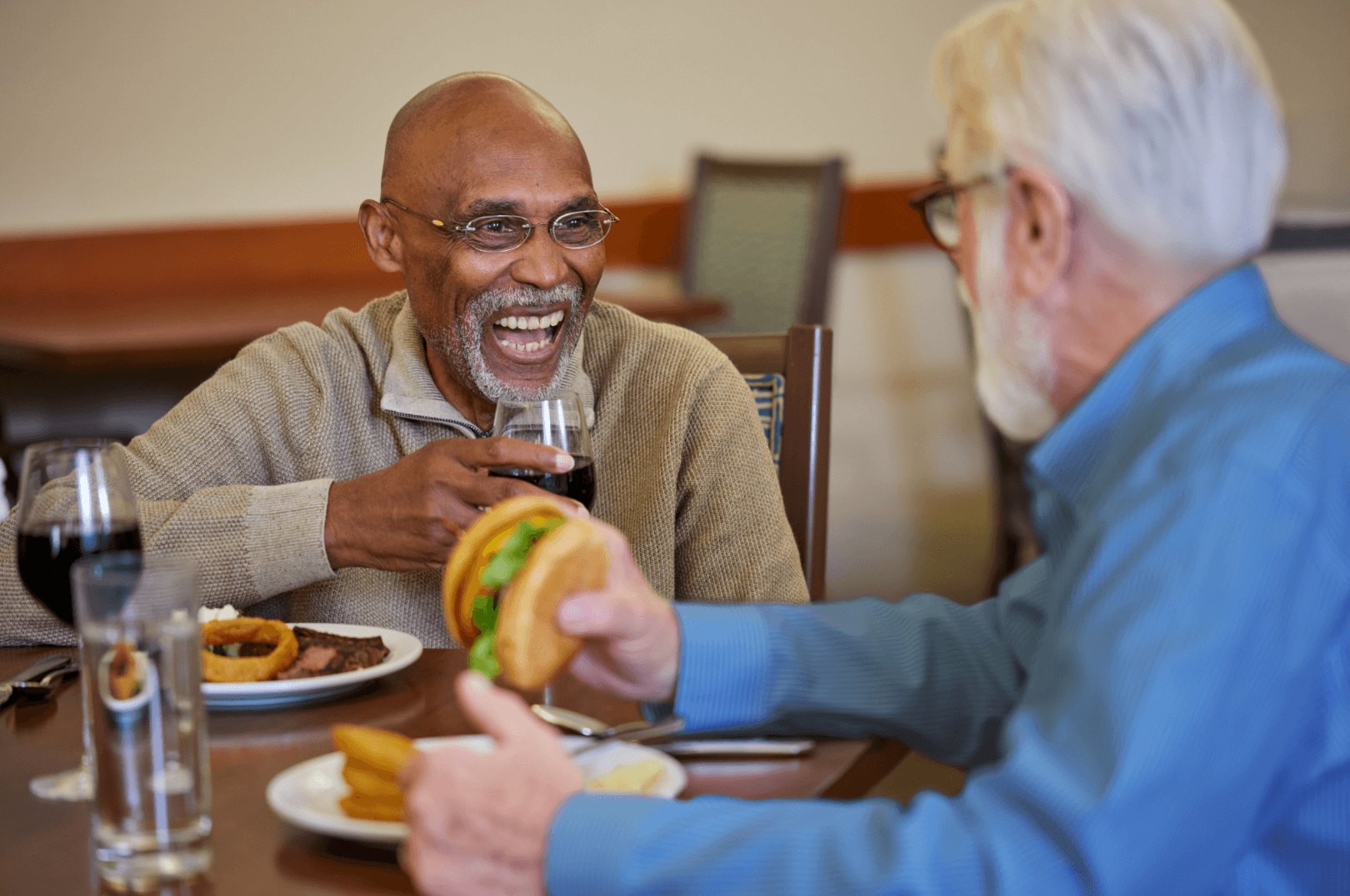 Two older men laugh and clink wine glasses over plates of food at an indoor dining table.