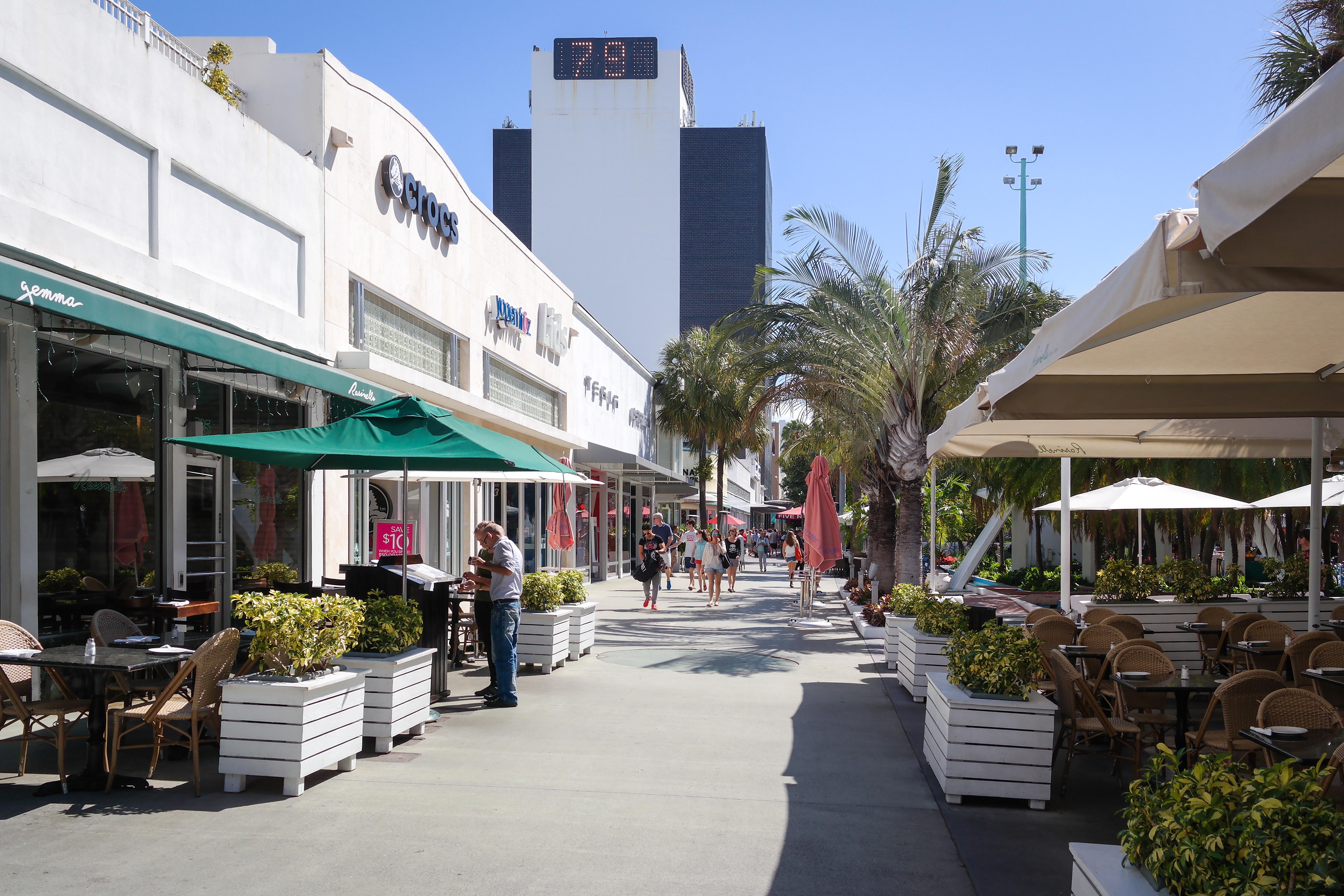 Wide pedestrian mall lined with outdoor dining, palm trees, and umbrellas under a blue sky.