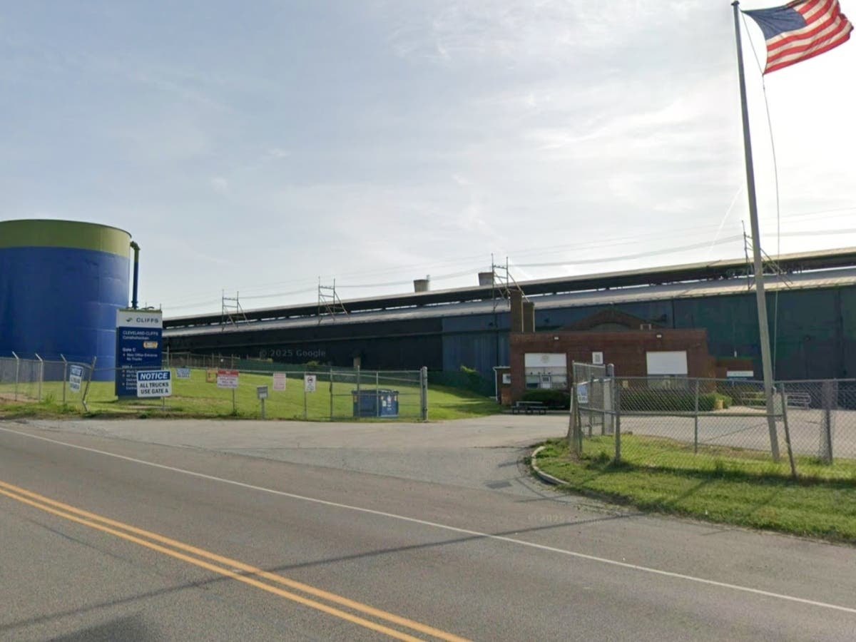 Large industrial building complex behind a chain-link fence with an American flag on a tall pole.
