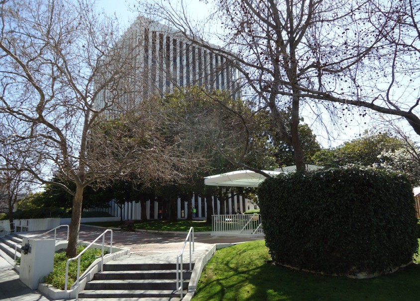 Modernist office building behind bare trees with a green lawn and hedges in the foreground.