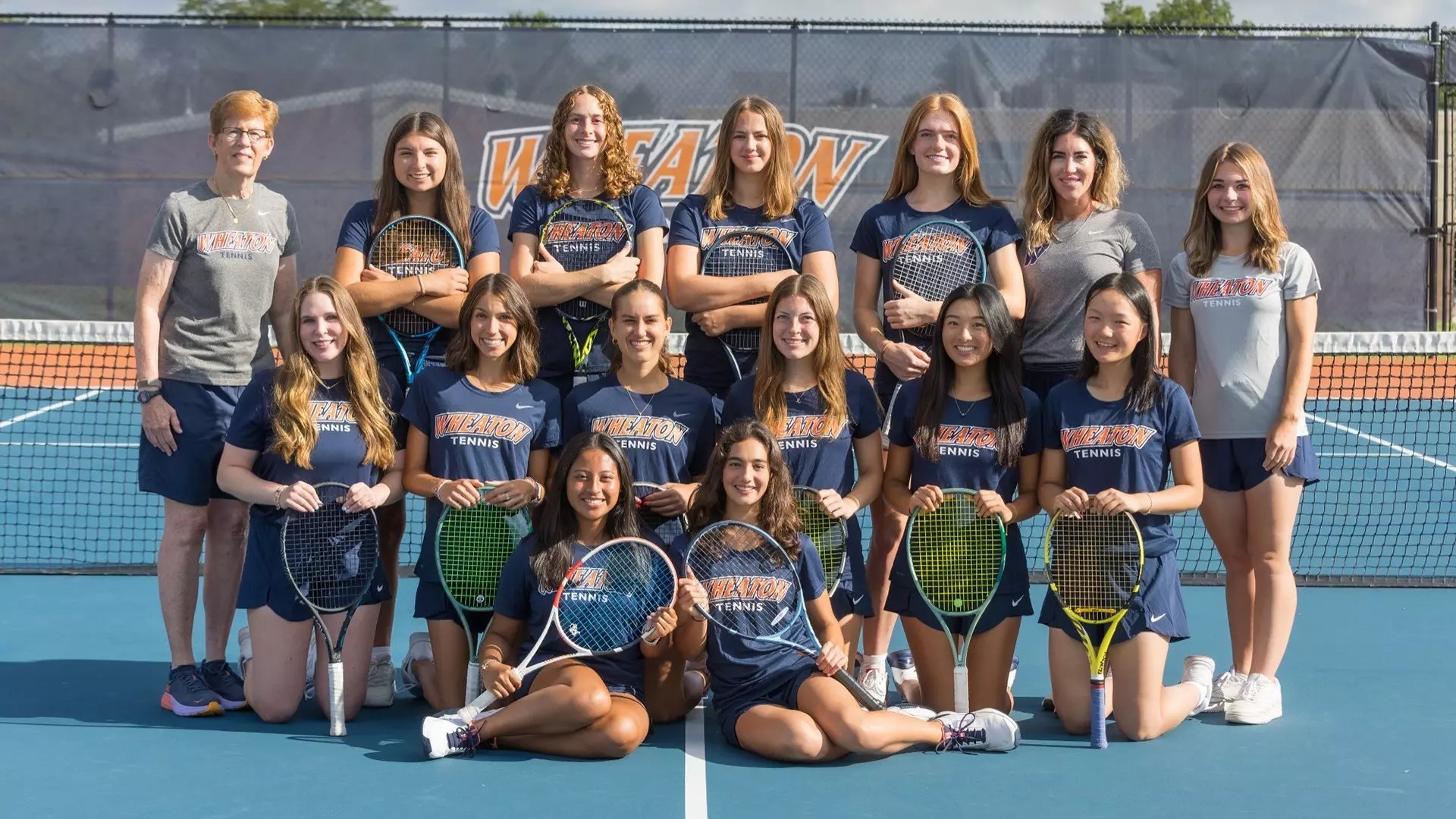 Group of women in navy Wheaton Tennis shirts posing on a blue and orange tennis court.