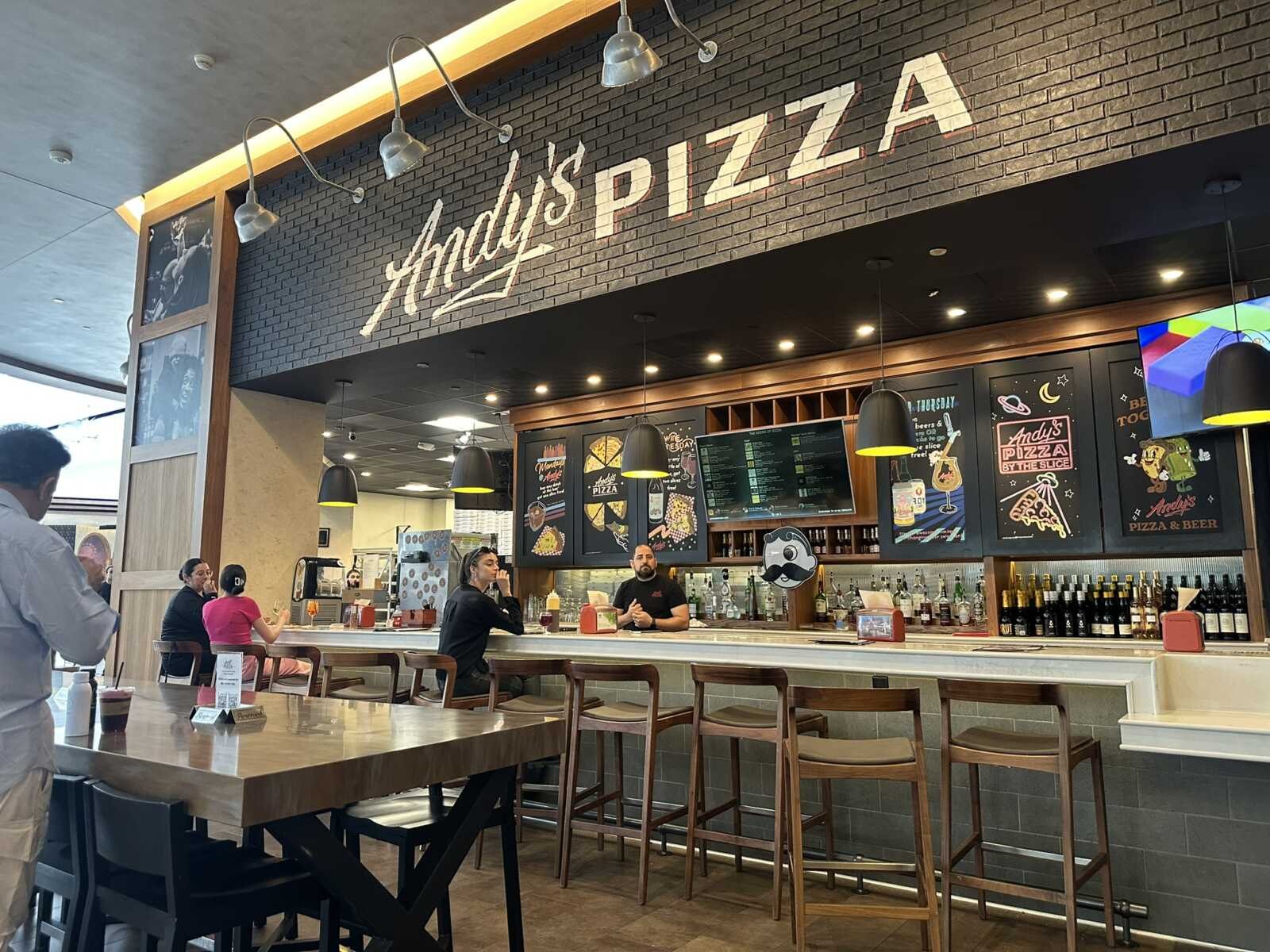 Interior of Andy's Pizza showing a counter with bar stools, brick wall with logo, and customers dining.
