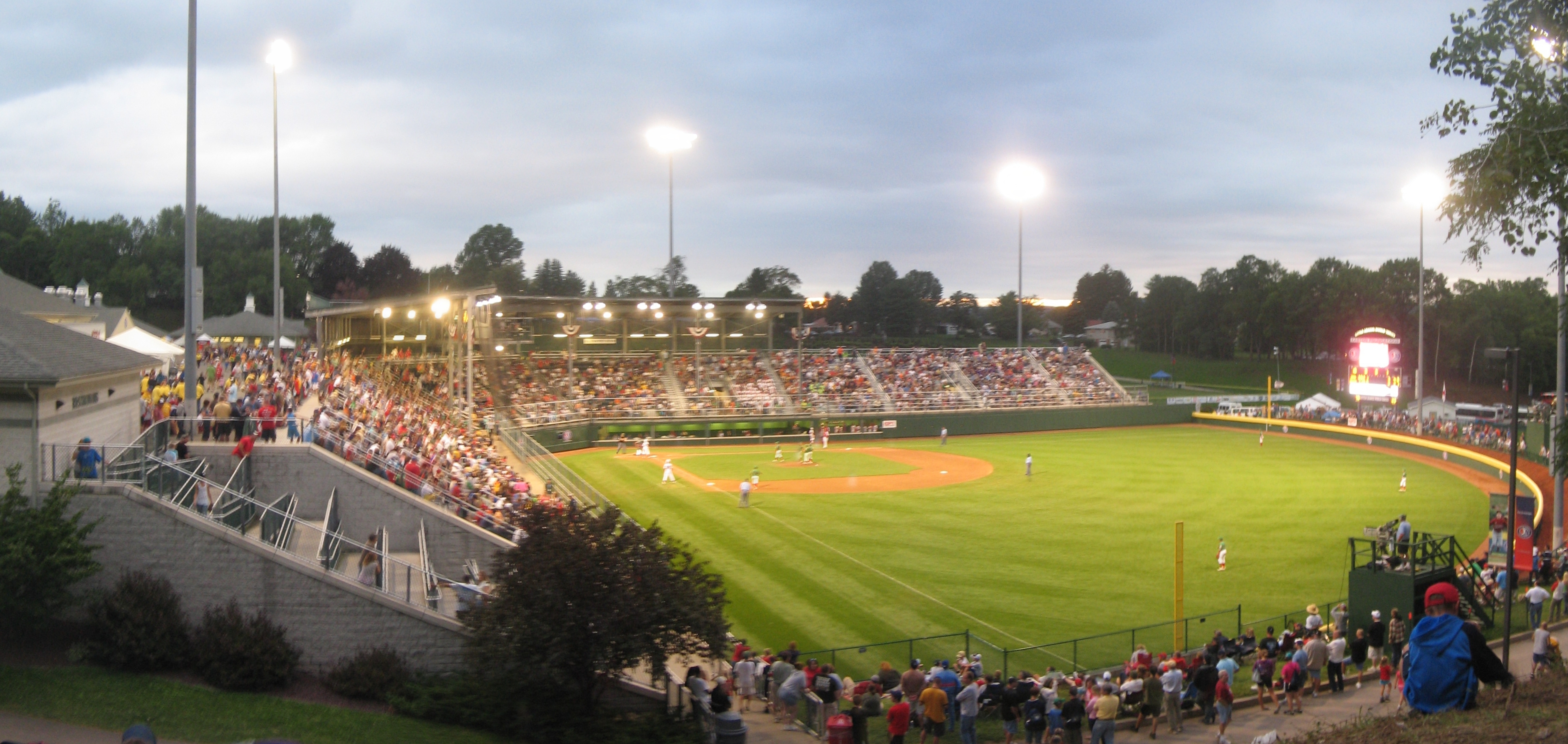 Panoramic view of a Little League stadium at dusk with lights on and packed stands.