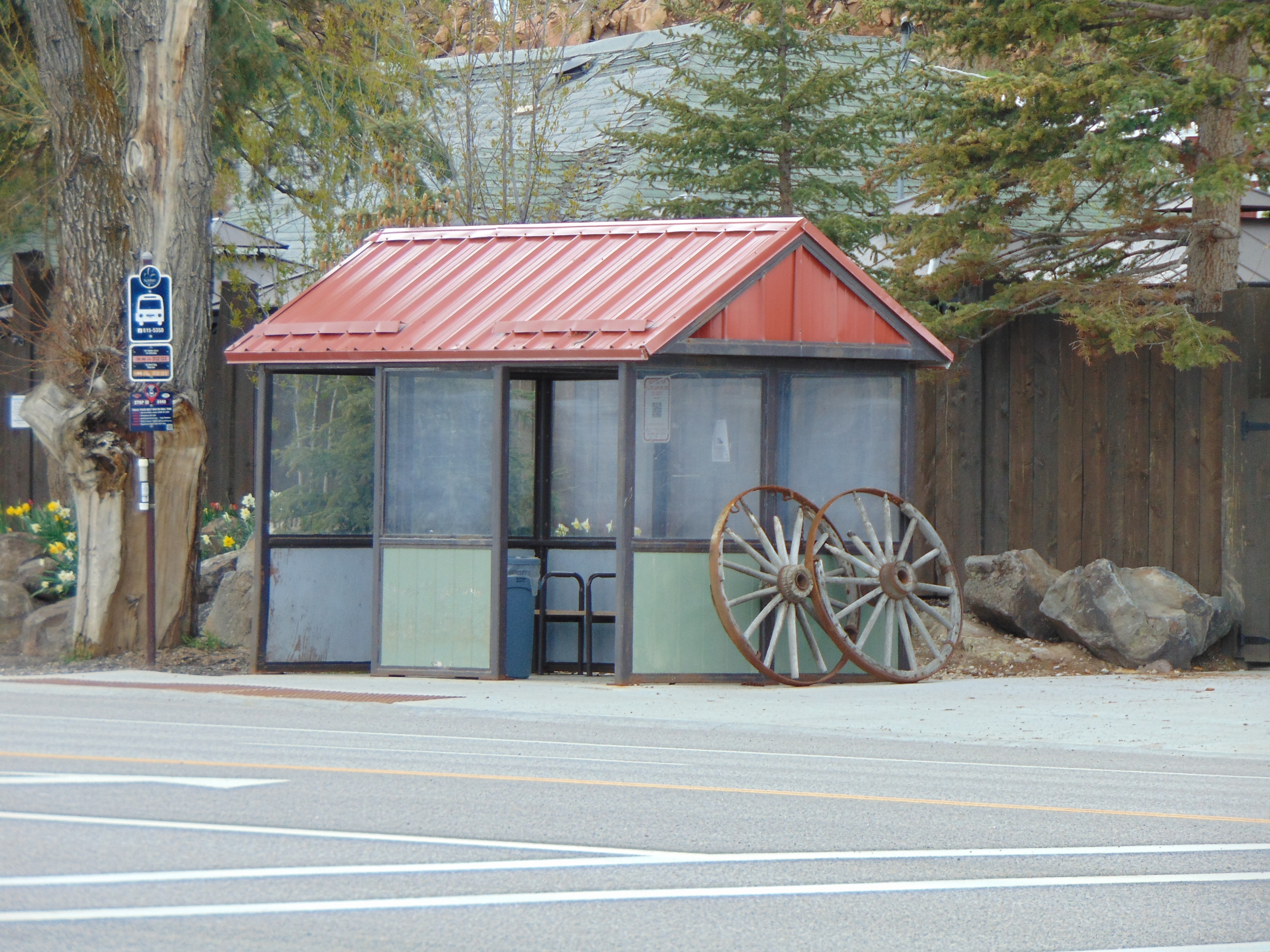 Glass-and-metal bus shelter with red roof along a road, wagon wheel decoration beside it.