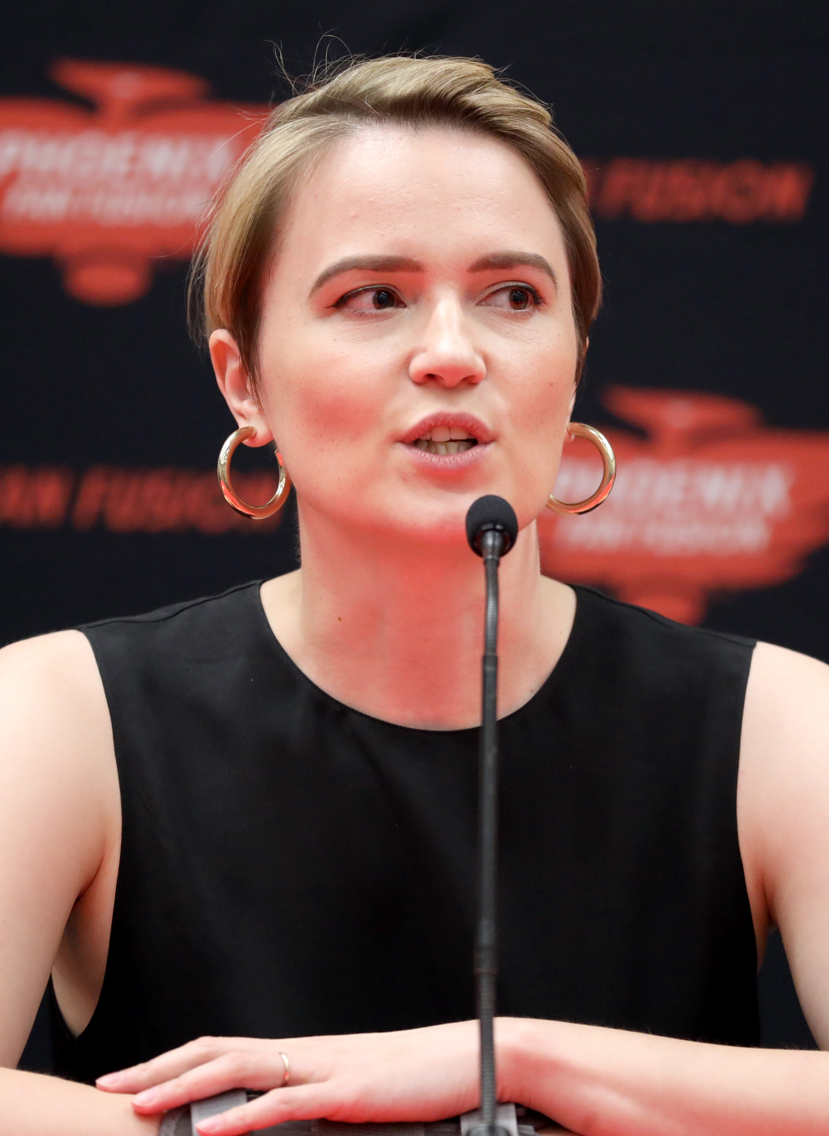 Woman with short blonde hair speaks at a microphone at a public event.
