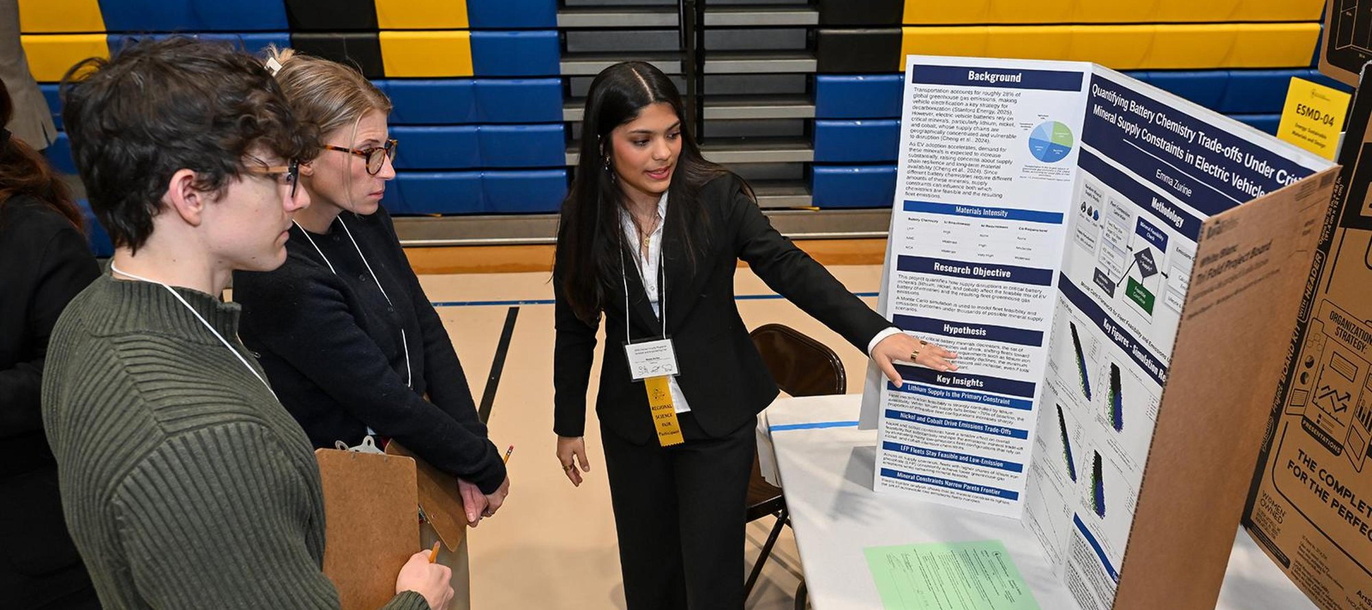 A student in a black blazer points at her research poster while two other students observe at a science fair in a gymnasium.