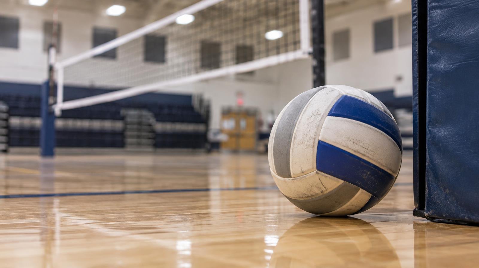 A volleyball resting on a hardwood floor near a volleyball net post.