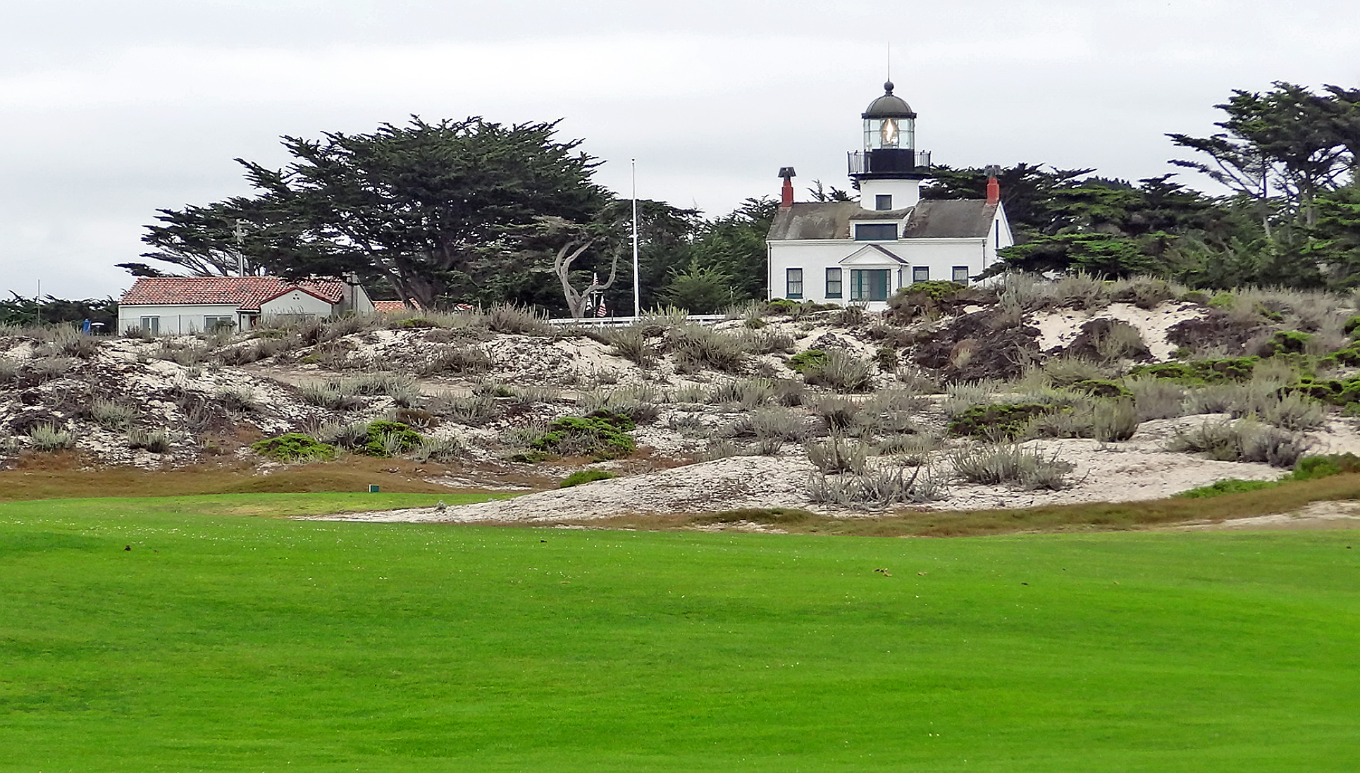 Coastal golf course fairway with a white lighthouse and cypress trees in the background.