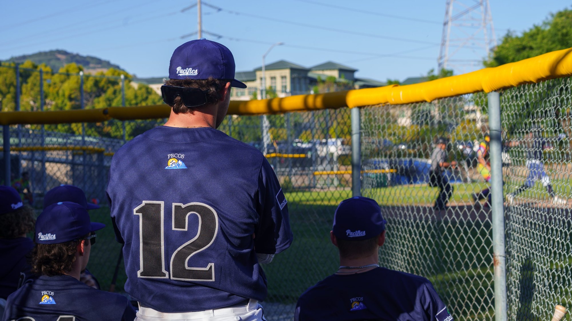 Baseball players in navy uniforms stand at a chain-link fence watching the field.