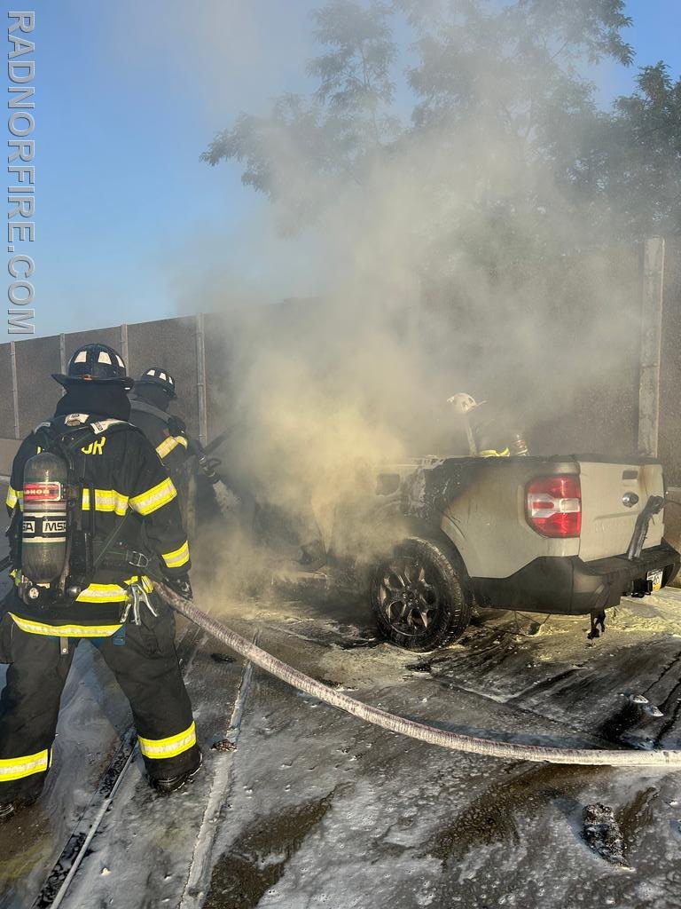 Firefighter in full gear spraying water on a burning pickup truck surrounded by heavy smoke on a highway ramp.