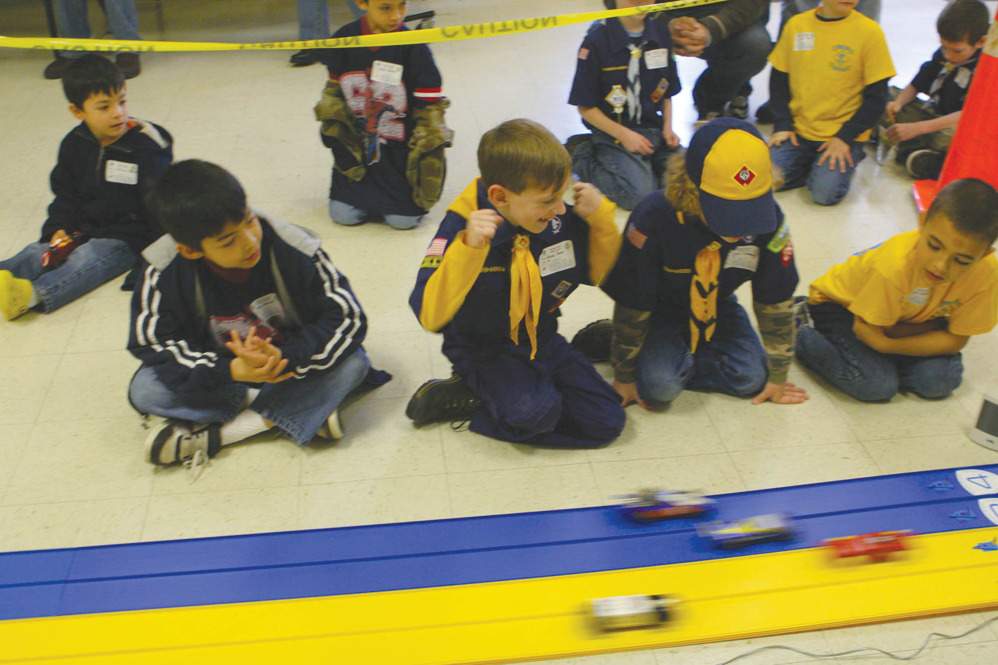 Children in Cub Scout uniforms crowd around a miniature wooden derby car racing track.