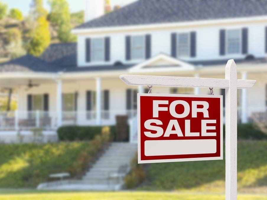 Red 'For Sale' sign on a post in front of a large two-story white house.