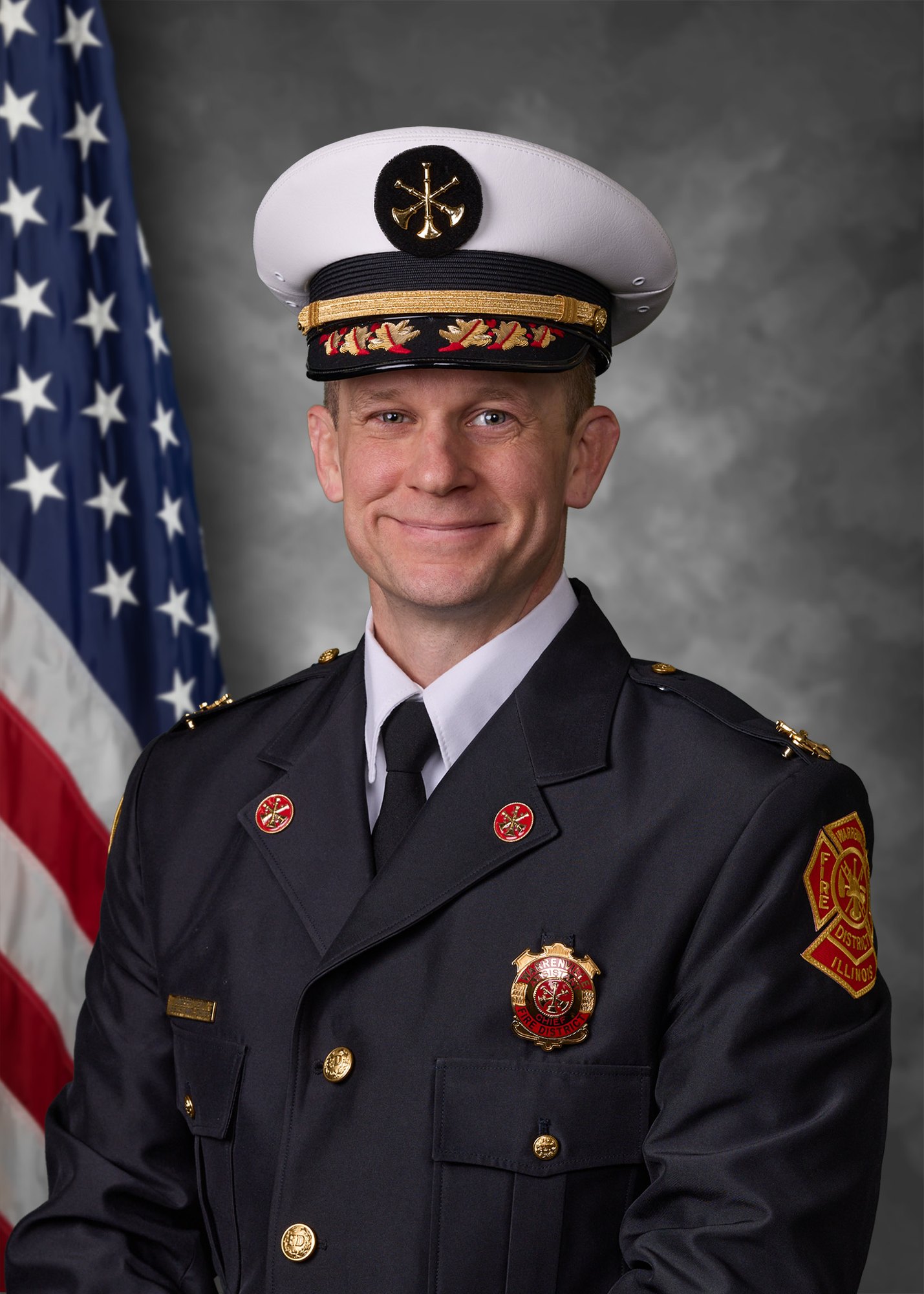 A man in a fire department dress uniform and white chief's cap posed in front of an American flag.