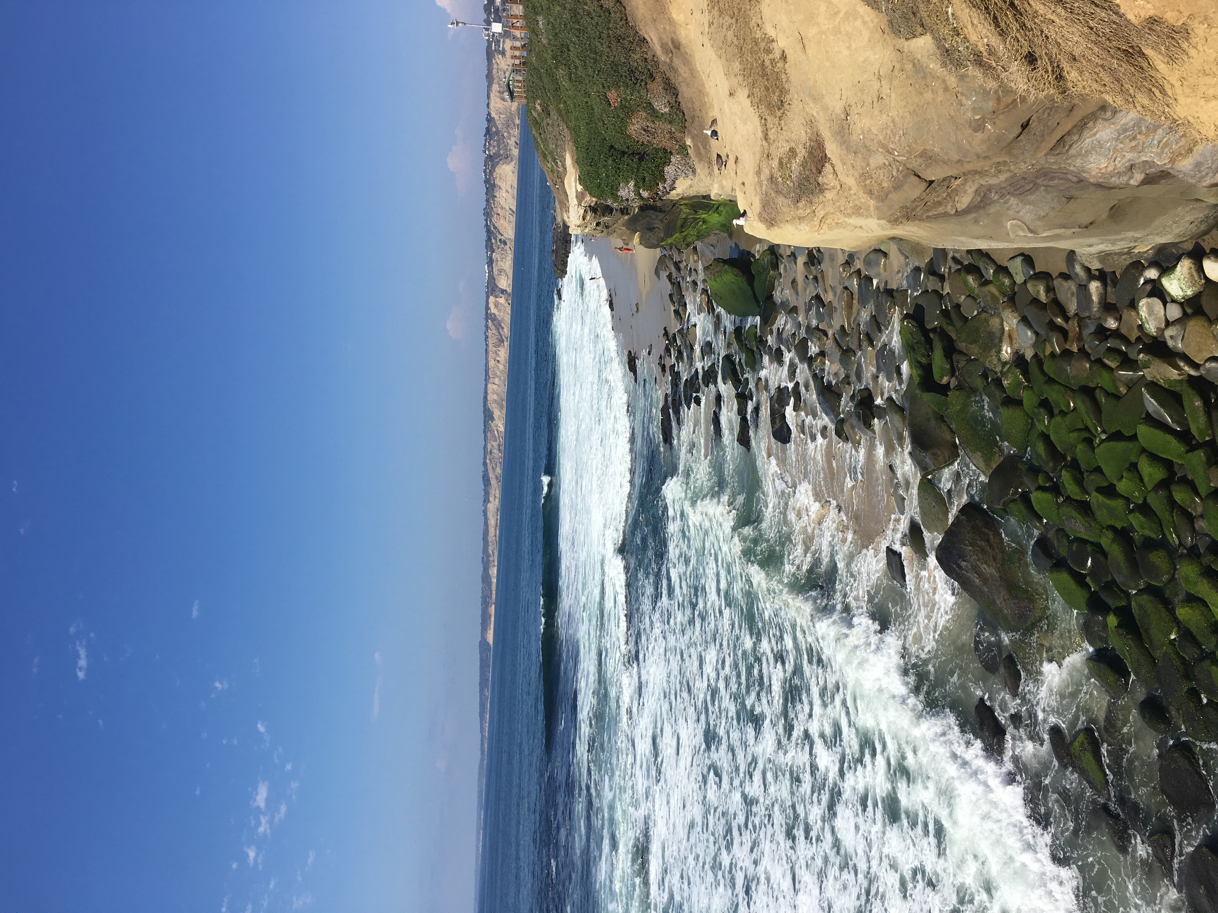 Rocky shoreline with algae-covered boulders and breaking ocean waves under a blue sky.