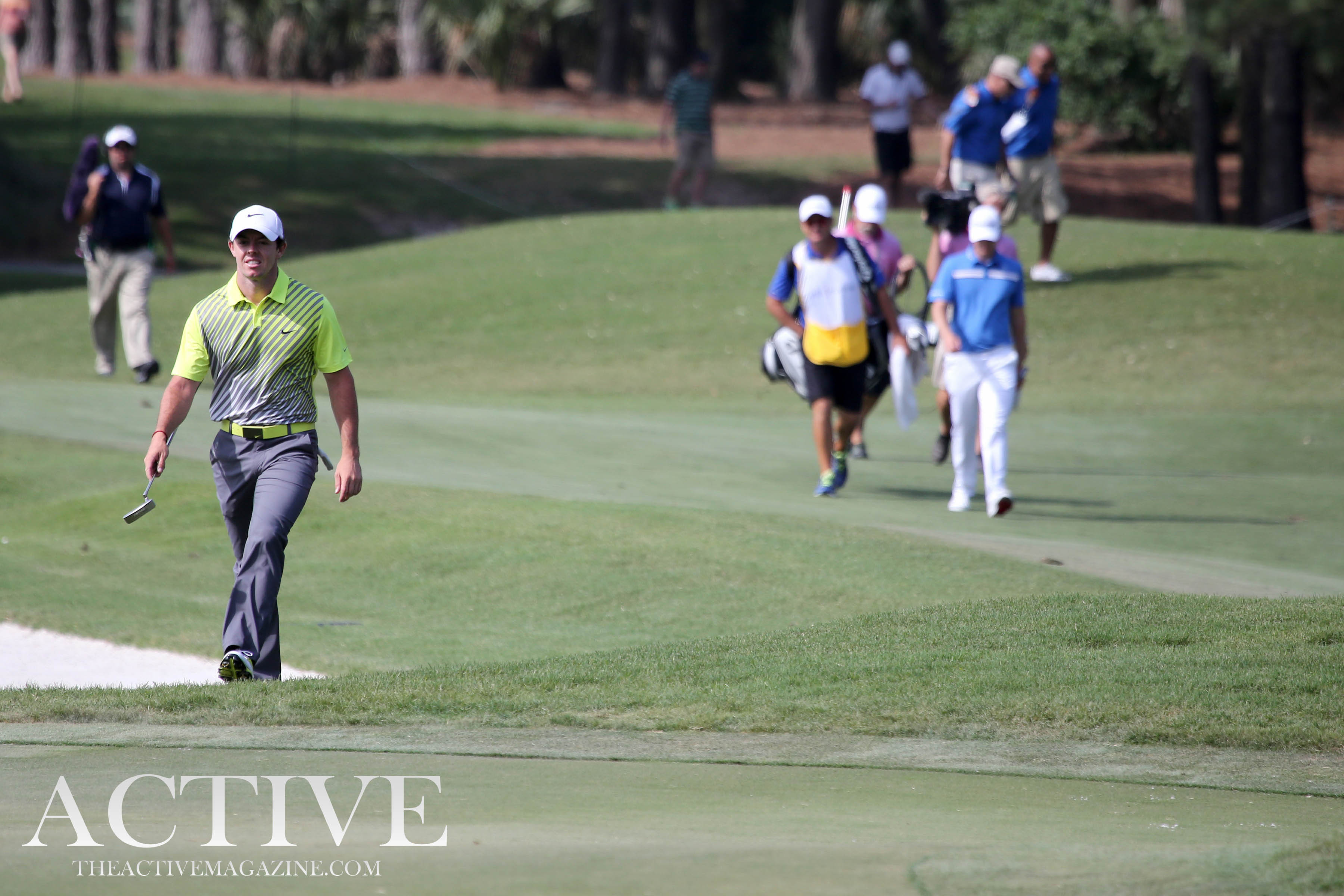 A golfer in a green polo walks a pine-lined fairway with caddies and trees in the background.