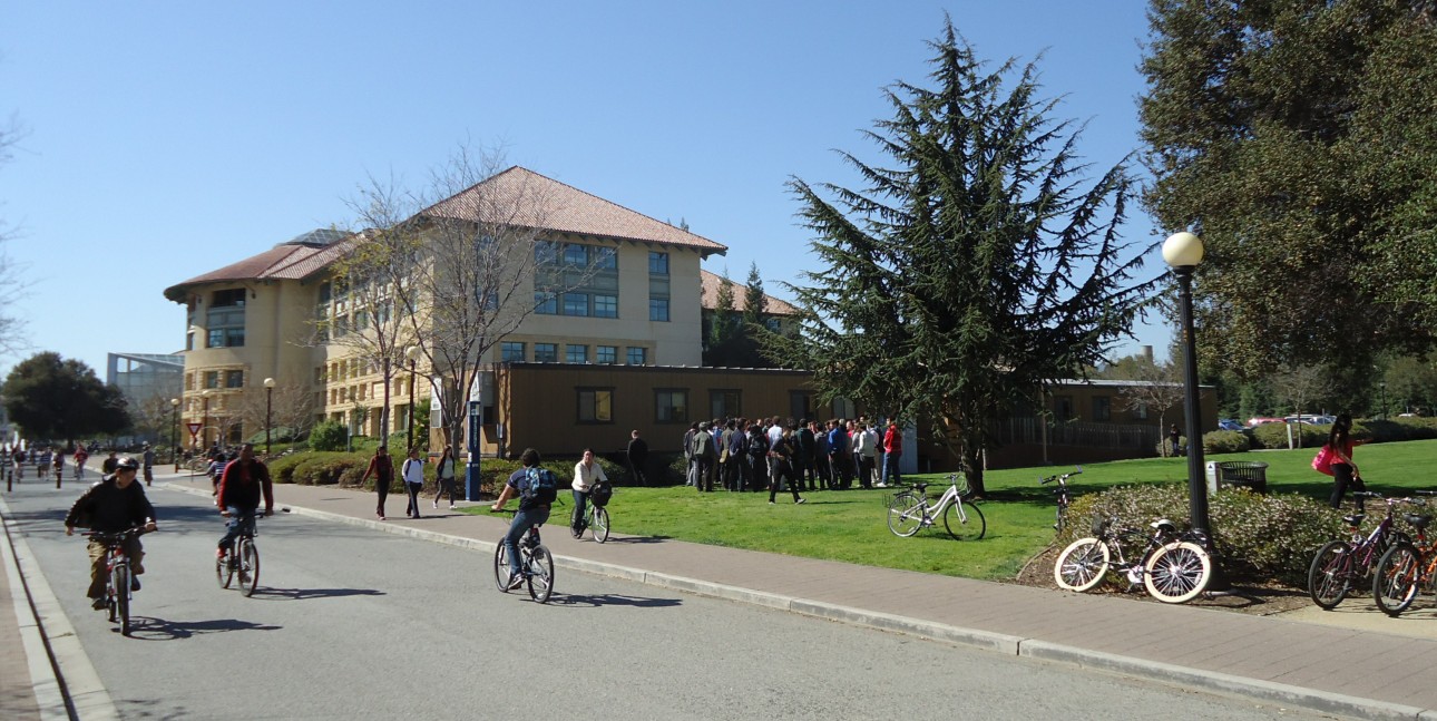 Students walking and cycling on a sunny university campus near multi-story academic buildings.