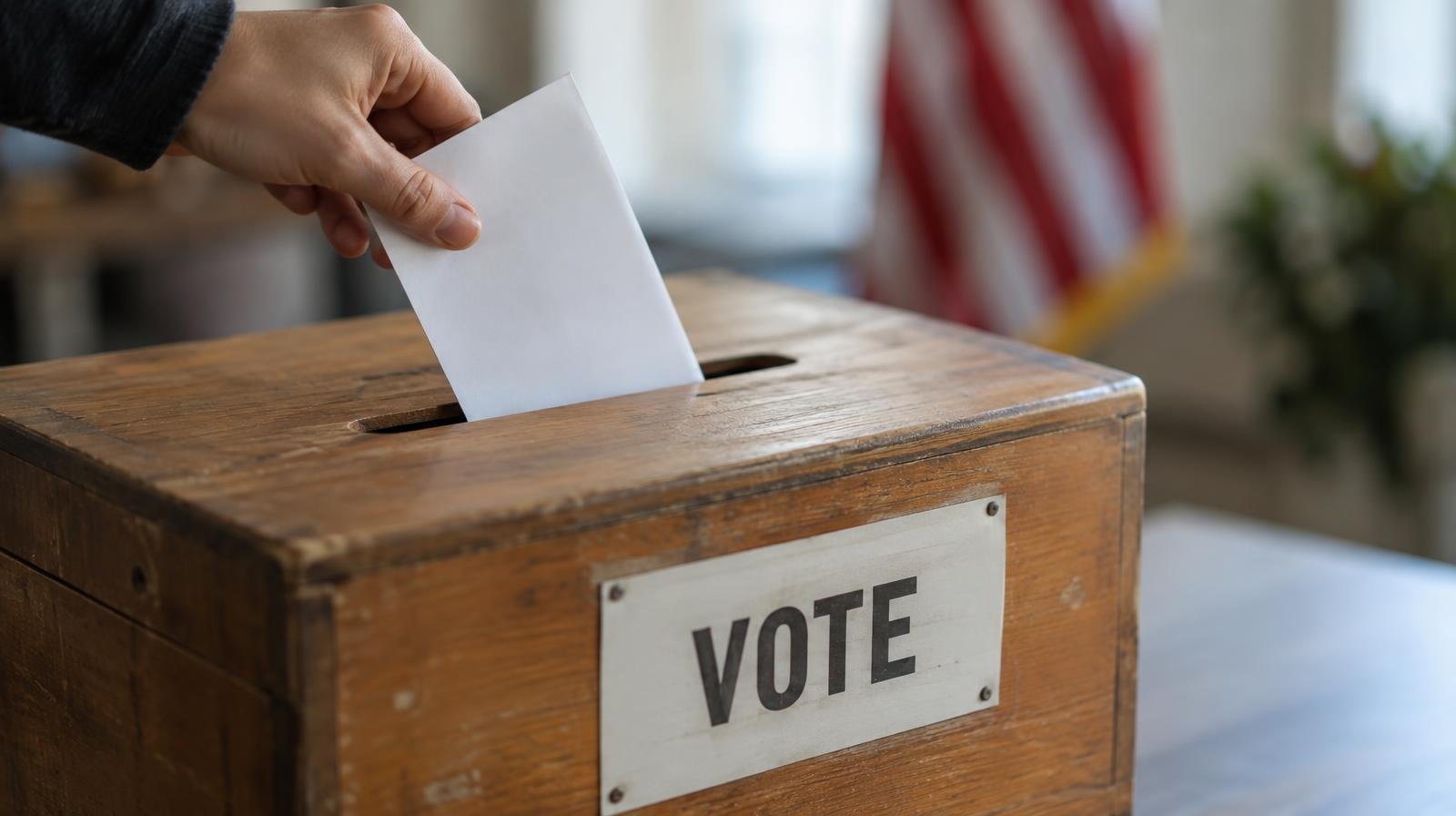 A wooden ballot box with a slot on top, sitting on a plain surface.