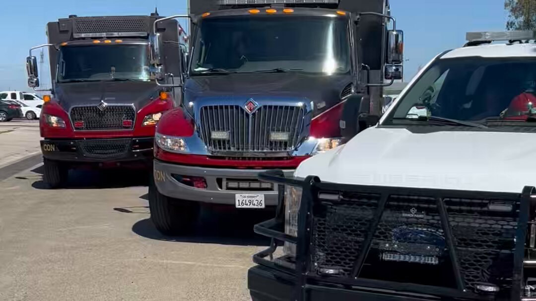 Three large red fire utility trucks parked in a sunny lot, with a white pickup truck alongside.