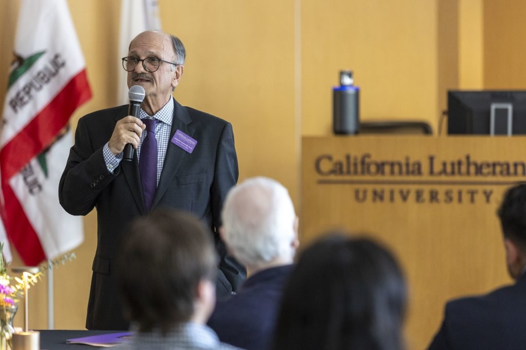 A man in a dark suit speaks at a podium bearing a university logo.