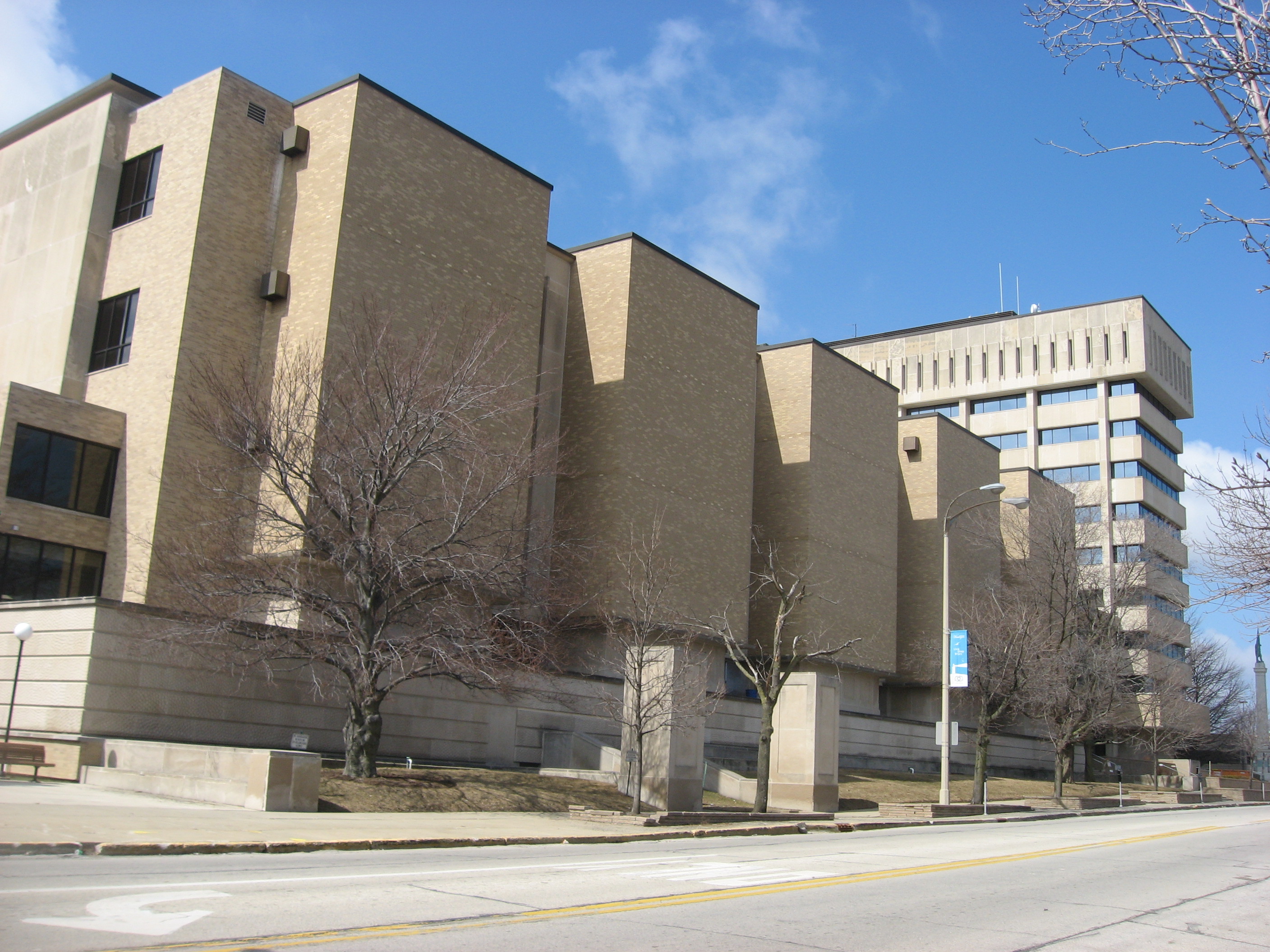 Large tan brick government building with bare trees along the sidewalk.