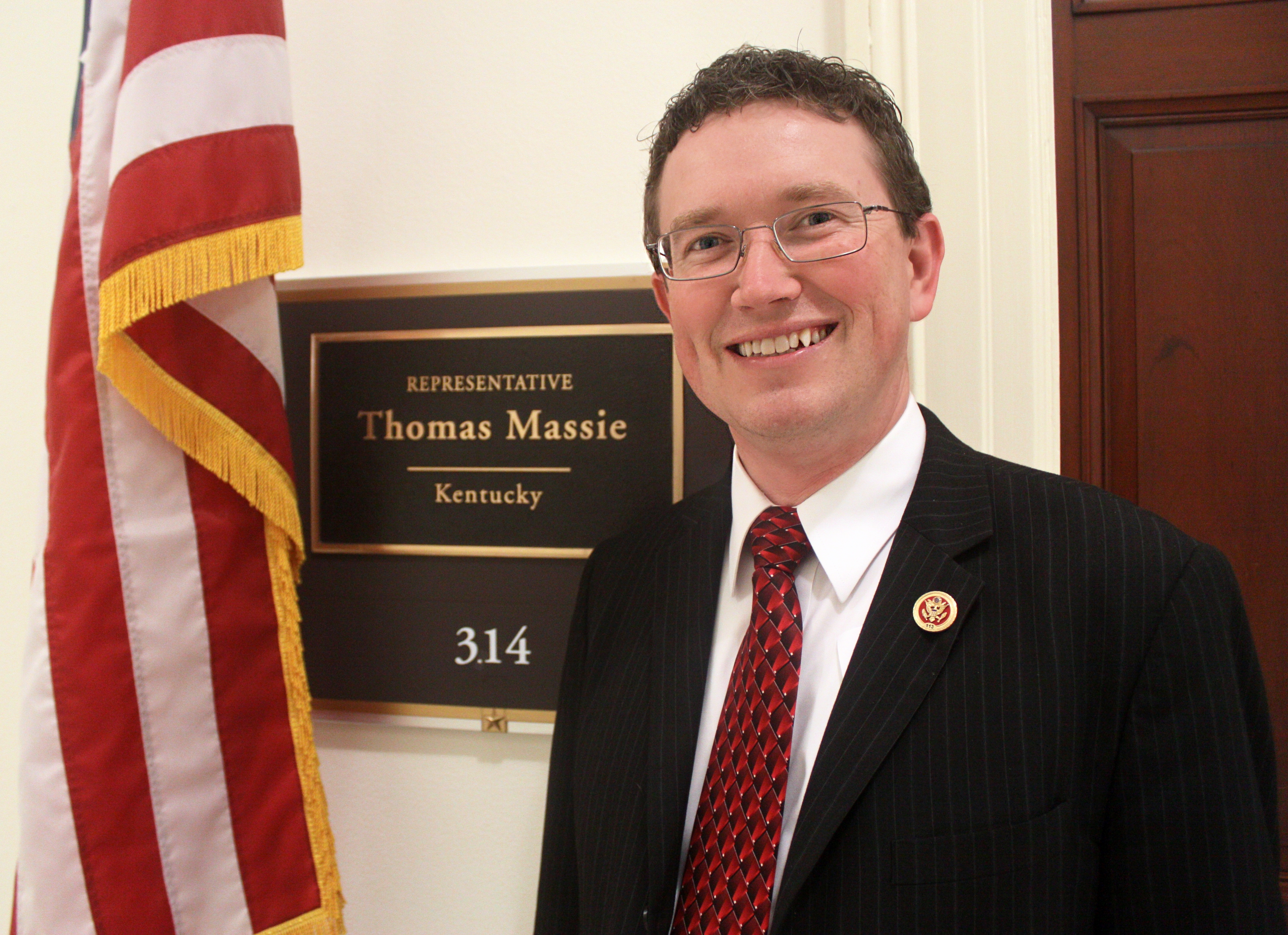 Man in dark suit standing next to a congressional office nameplate with an American flag behind him.
