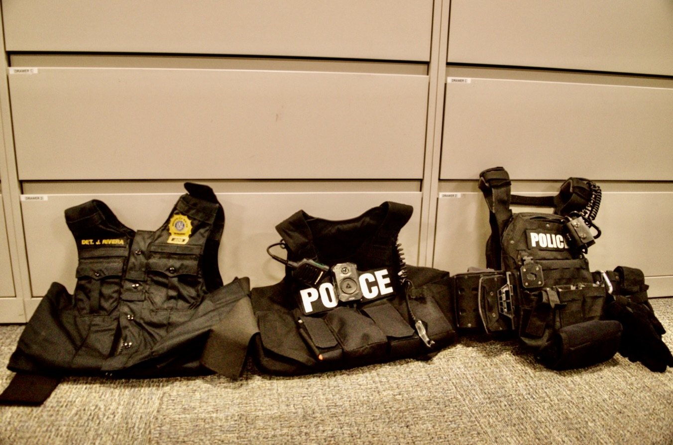 Three black ballistic vests with police markings and tactical gear displayed side by side against a wall.
