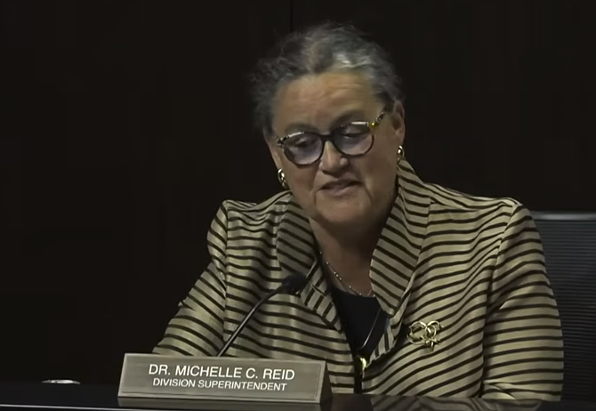 A woman with gray hair and glasses in a striped blazer speaks at a meeting dais with a nameplate.