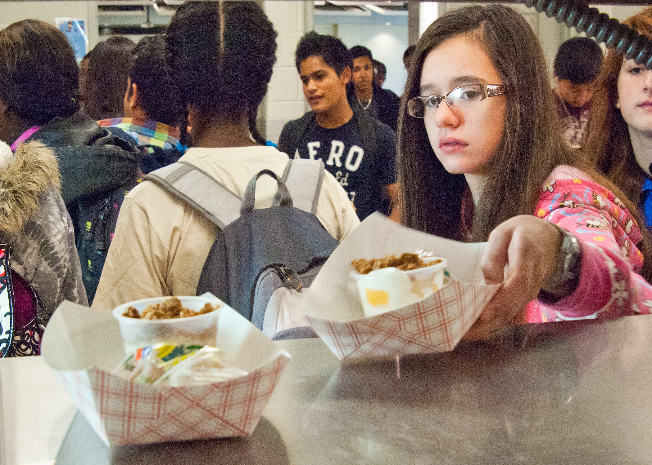 Students holding food trays in a school cafeteria serving line.
