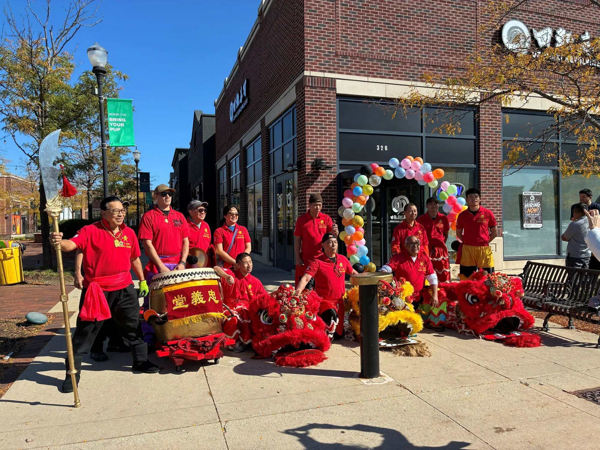 Group of performers in red costumes with lion dance props posed outside a brick storefront with a balloon arch.