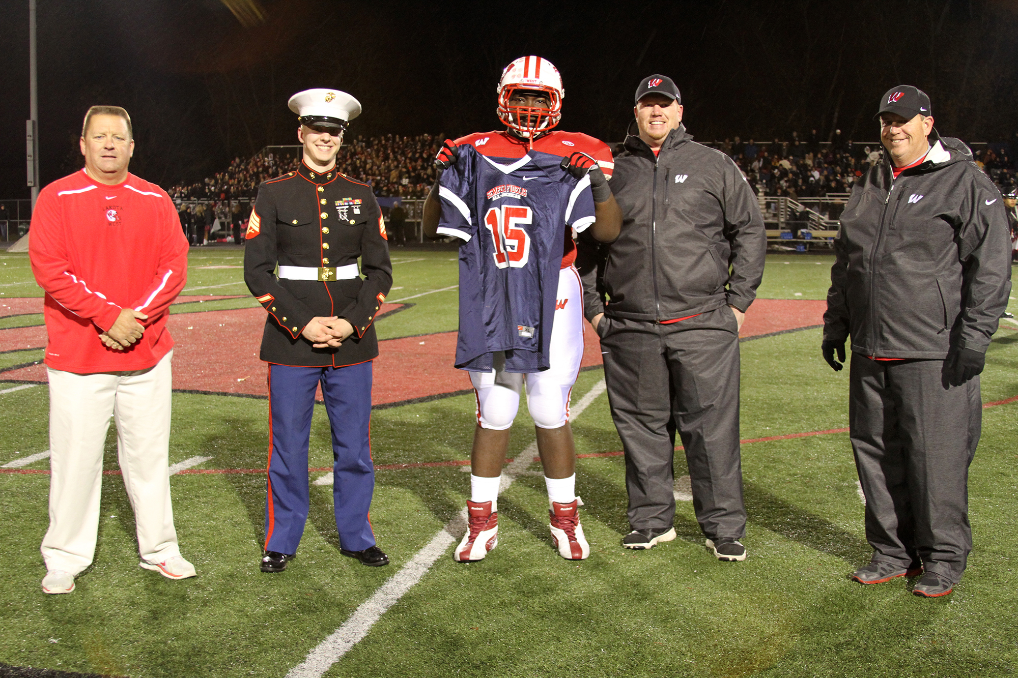 Football player in red and white uniform holds a jersey at a nighttime on-field ceremony.