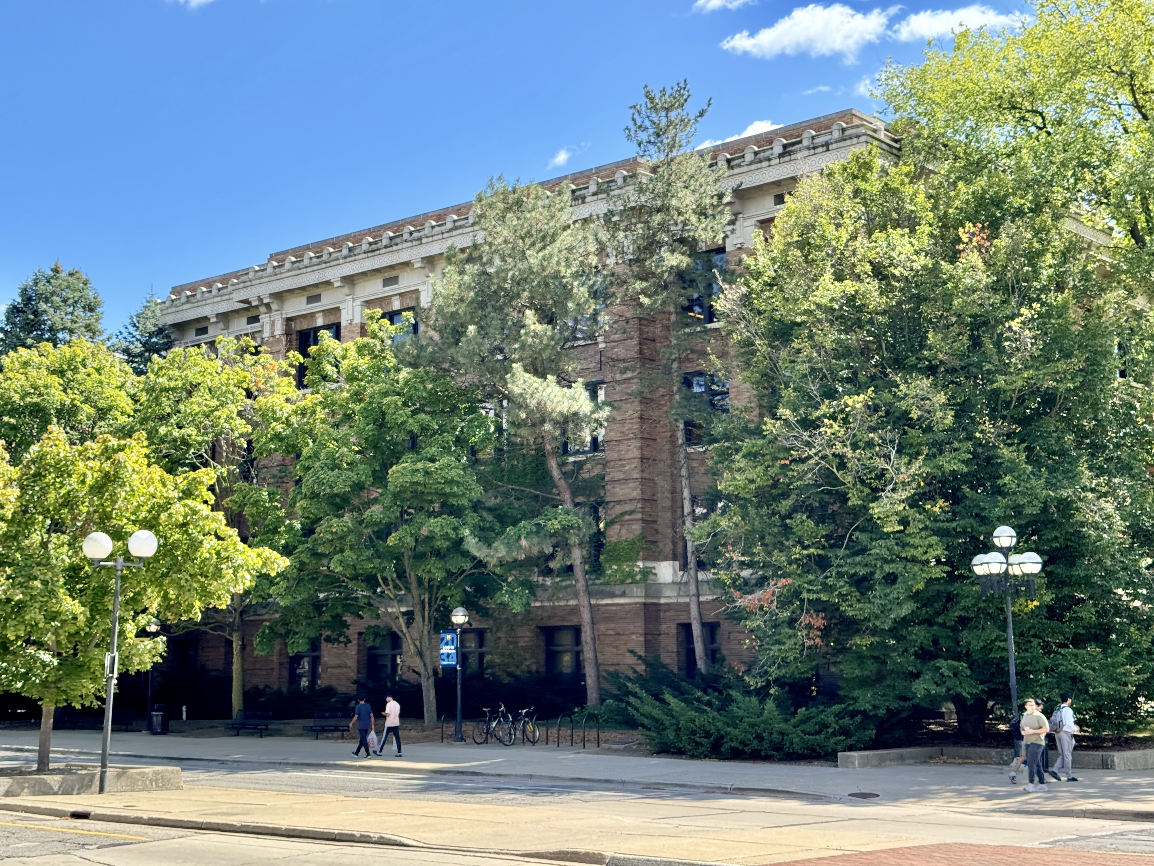 Brick university building partially obscured by trees, with bicycle racks and pedestrians in front.