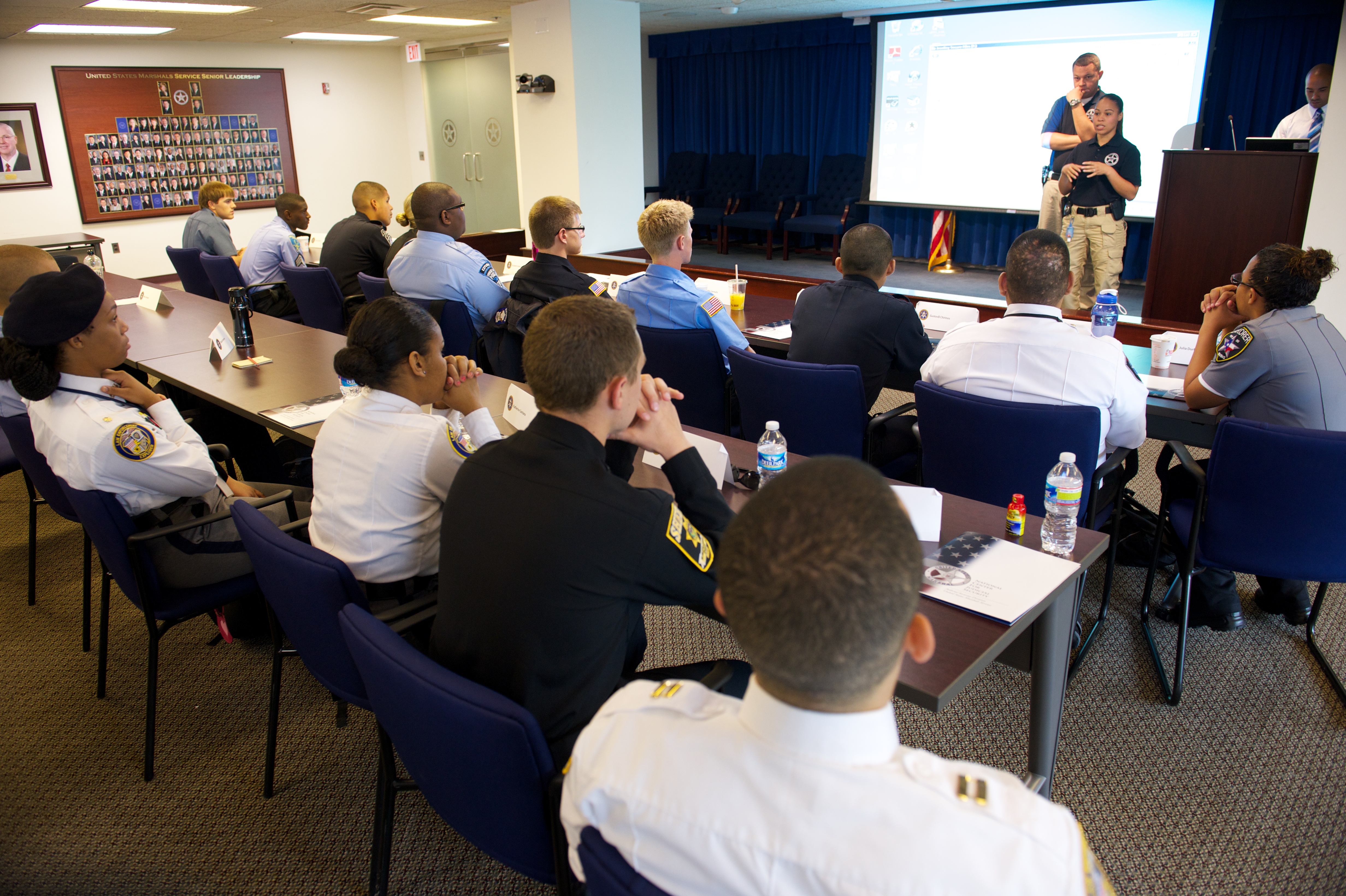 Rows of uniformed officers and civilians seated in a conference room watching a presenter.