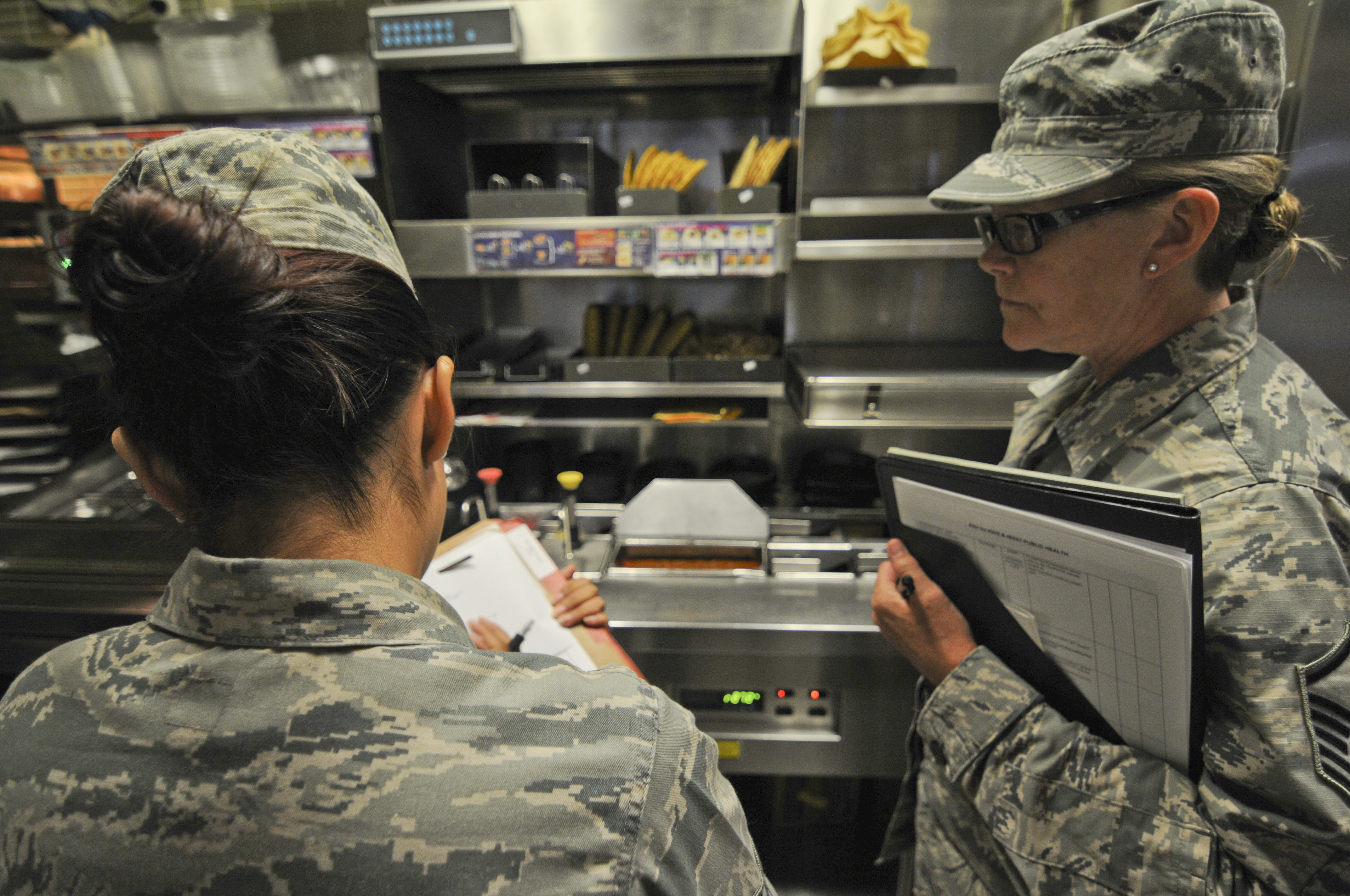 Two uniformed inspectors holding clipboards review food safety conditions in a commercial kitchen.