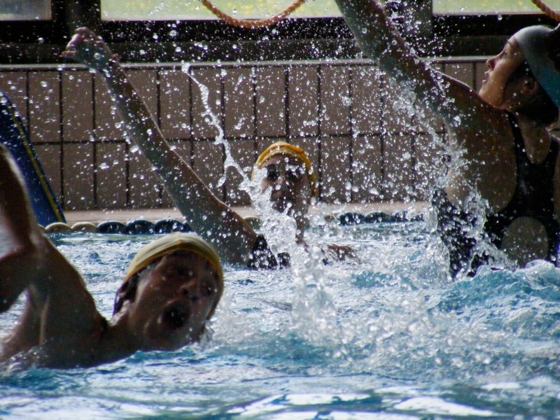 Multiple water polo players in swim caps compete in a pool with water splashing during active play.